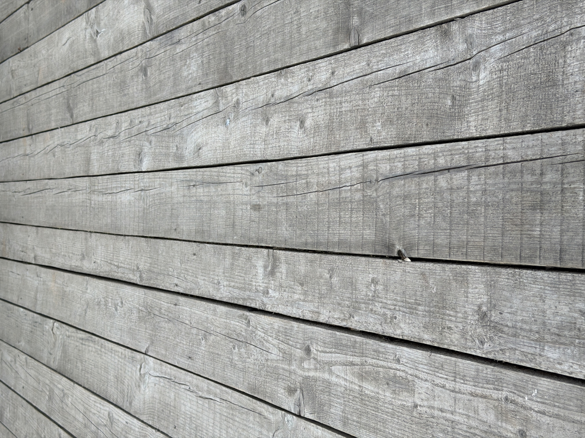 The image displays a close-up, detailed view of a weathered wooden wall. The wall is constructed from horizontal wooden planks, laid one above the other. The wood is gray in color and exhibits a rough texture, with visible grain lines, knots, and cracks, indicating age and exposure to the elements. The planks are not perfectly smooth; some have slight unevenness and wear.
The lighting suggests it is daytime, with sunlight casting subtle shadows and highlighting the texture of the wood. The perspective is slightly angled, giving a sense of depth and showing the continuity of the planks across the surface. There are no people or specific objects clearly identifiable in the scene, focusing the attention entirely on the material and construction of the wall. The setting appears to be outdoors, given the natural weathering of the wood. The overall impression is one of rustic simplicity and durability. No text is visible in the image.
