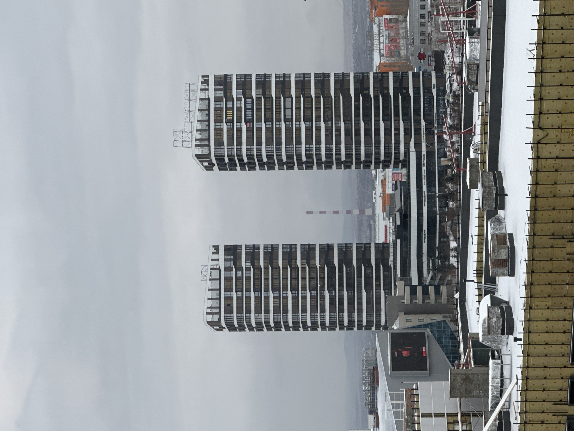 The image presents a view in Krasnoyarsk, Russia, likely from a high vantage point. The primary subjects are two identical high-rise buildings with a distinct modern architectural style, characterized by evenly spaced vertical columns and numerous windows. A muted color palette is dominant.
Foreground elements include what appears to be a construction or rooftop area covered in snow, with construction materials and structural elements visible. There are also red-colored rods.
In the background, the skyline shows other buildings of varying heights, indicative of an urban setting, further back there is smoke rising. The sky is overcast, lending a gray, somewhat bleak atmosphere to the scene. Overall, the image captures a cold urban environment, possibly in a developing area.