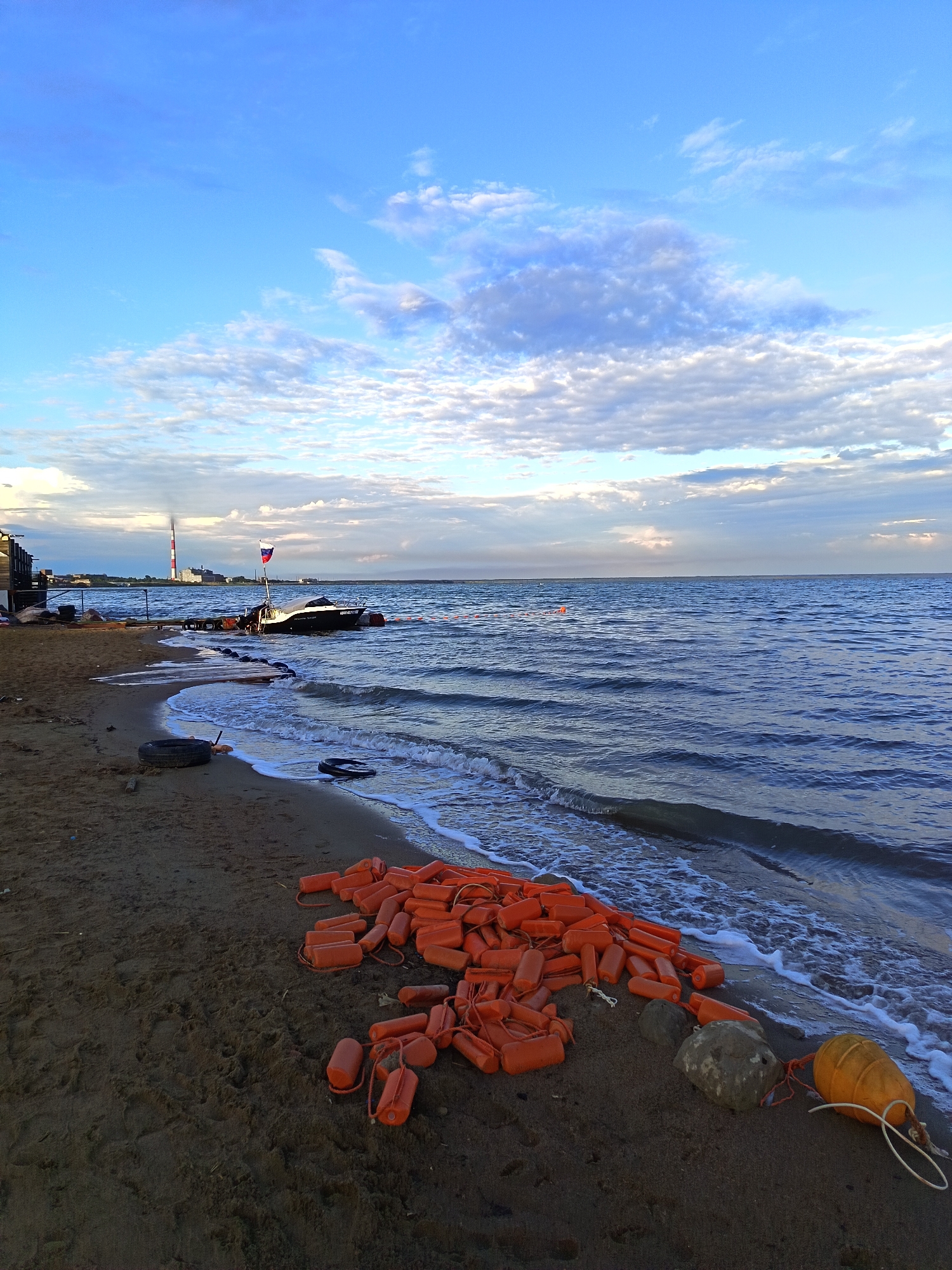 This is a wide-angle, eye-level photograph taken on a sandy beach in Yarovoye, Russia, facing a body of water. The sky is a vibrant blue with scattered white and gray clouds. The water, a deep blue, is gently lapping at the shore, creating white foam.
On the beach, in the foreground and mid-ground, a large pile of bright orange buoys is spread out. Some of these buoys are connected by rope. There are also a few large rocks and what appear to be tire remnants scattered on the sand. The sand itself is dark and appears wet near the water's edge.
In the water, a dark boat with a Russian flag is anchored or tied to a wooden pier. Further in the distance, a tall, red and white striped smokestack is visible, indicating industrial activity. To the left of the smokestack, a low-rise building or structure is present. The overall scene suggests a calm, somewhat industrial coastal environment. There are no people visible in the photograph.