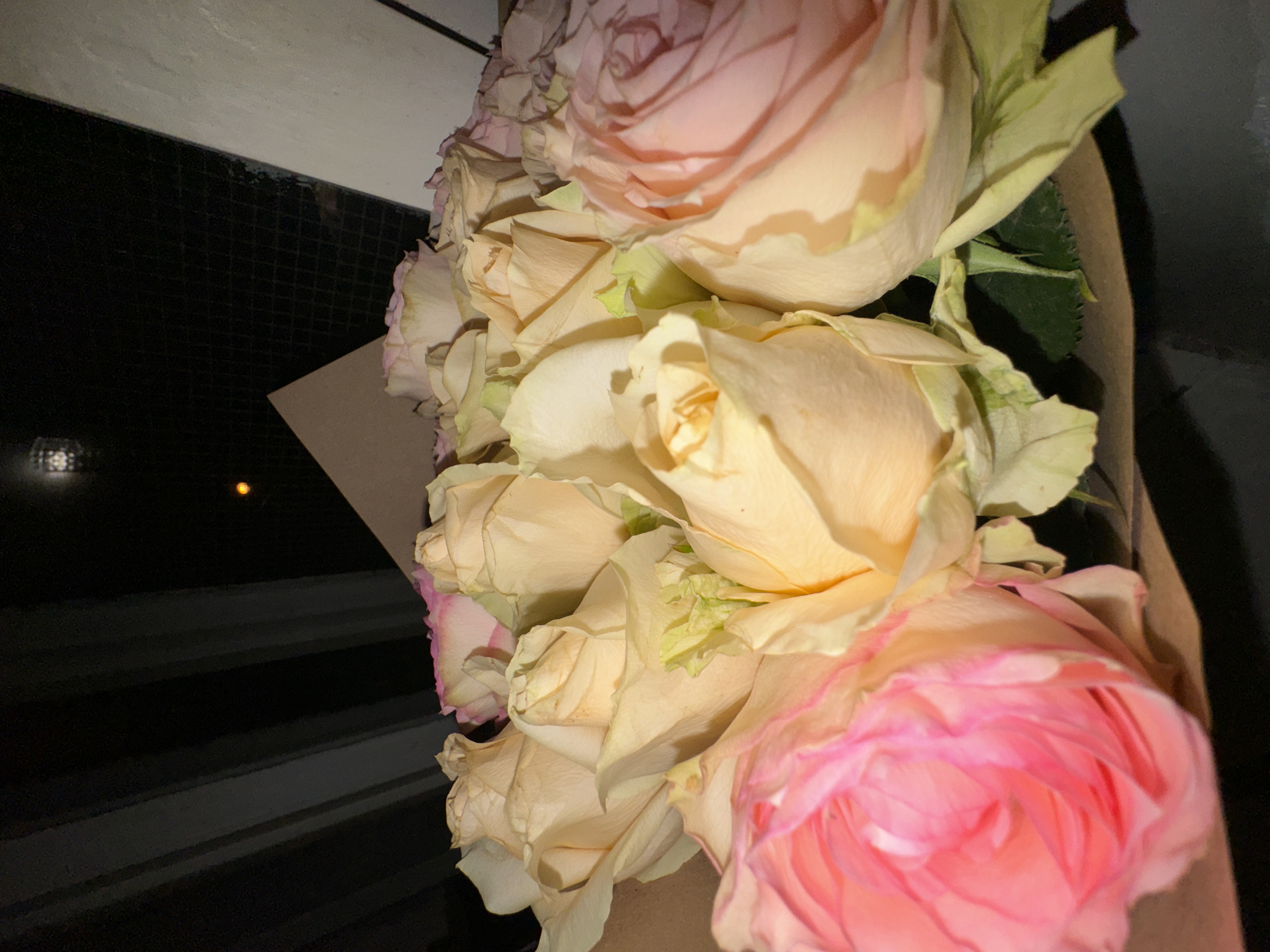 A bouquet of roses, predominantly pink and peach in color, is positioned in front of what appears to be a window or reflective surface, possibly in Moscow, Russia. The flowers are partially wrapped in brown paper. The background features a dark, possibly black, linear pattern. A single, small yellow light reflection is visible on the left side of the image.