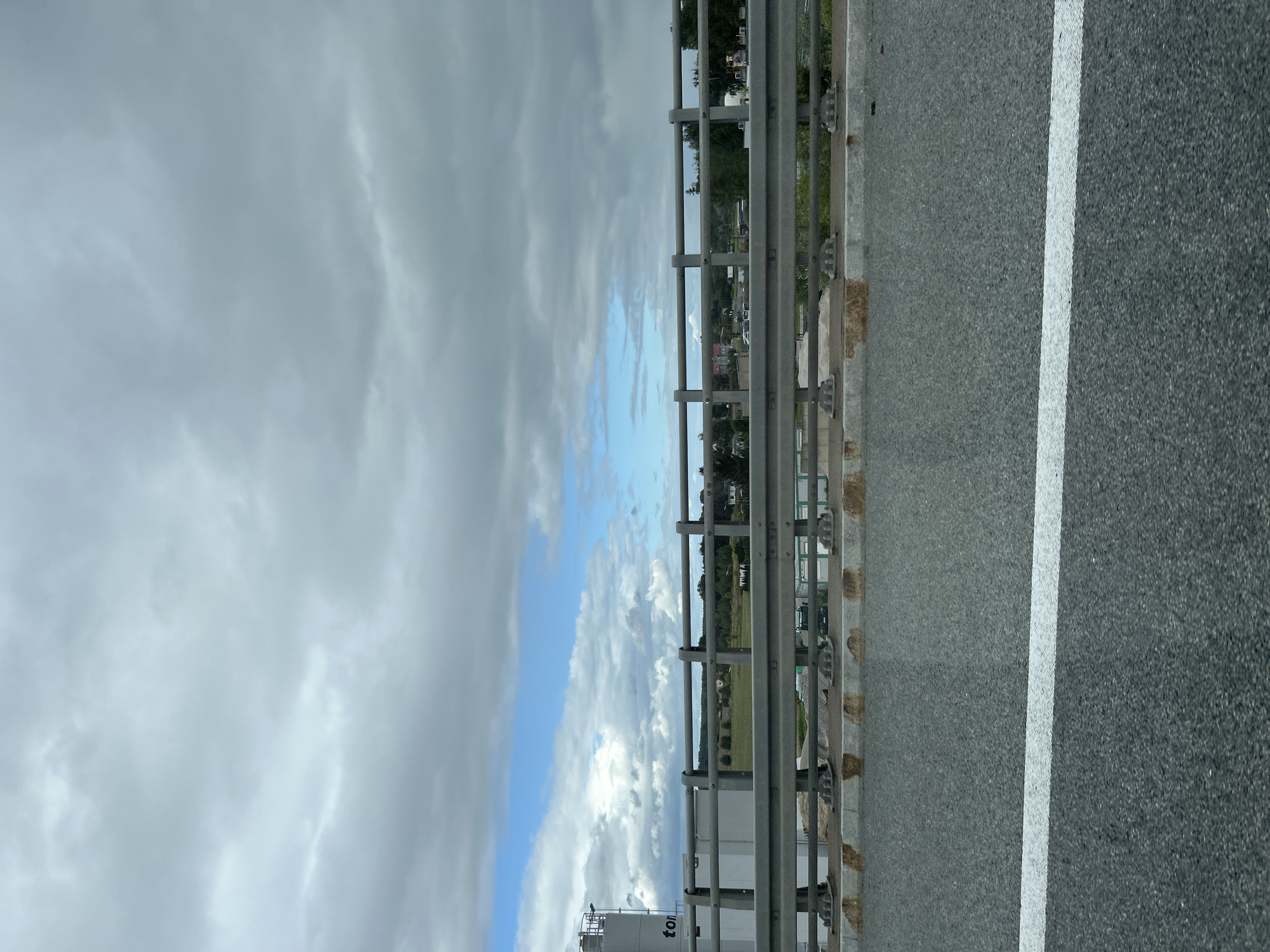 The image shows a view from a highway bridge over a rural landscape. In the foreground, a white line marks a lane on the asphalt road. Beyond the road, a metal guardrail runs horizontally across the frame, with rusty stains visible on the concrete base. In the background, a green field stretches to the horizon, dotted with trees and some buildings. A large industrial silo with the word "ton" visible on it stands to the left. The sky is filled with gray clouds with patches of blue sky visible. The location is identified as Świebodzin, Poland.