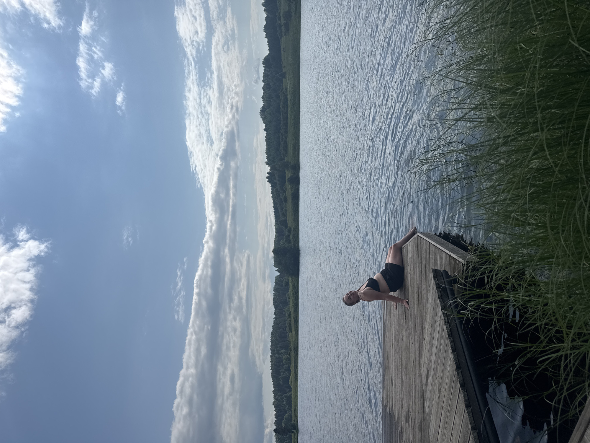 A woman in a black bikini top and black shorts is sitting on a wooden dock, facing the right side of the frame, with her legs extended. She is looking towards the camera with a slight smile. The dock is made of weathered wooden planks and extends into a body of water. The water is calm with ripples and reflects the blue sky. The background features a dense line of trees and vegetation, with a few scattered structures visible. The sky is blue with scattered white clouds. The overall scene suggests a peaceful summer day by a lake or river, possibly in a rural area. The location is identified as Pozdyshki, Russia.