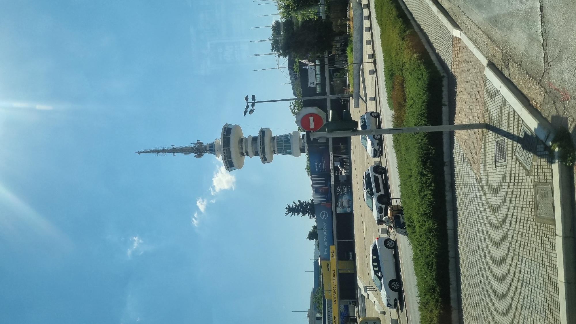 The image captures a daytime scene in the city of Thessaloniki Municipal Unit, Greece, featuring the iconic Thessaloniki Tower. The tower, a prominent landmark, stands tall in the background against a bright blue sky with scattered white clouds. Below the tower, a modern building complex is visible, with several cars parked in front and driving on a road. A white car, a white Citroën C4 Cactus, and a silver Citroën C1 are present. A person is riding a scooter, carrying a large box on the back. In the foreground, a neatly trimmed green hedge runs along a sidewalk. A traffic light with a red stop sign is also visible, indicating a street intersection. The overall ambiance is one of an urban environment on a clear day.