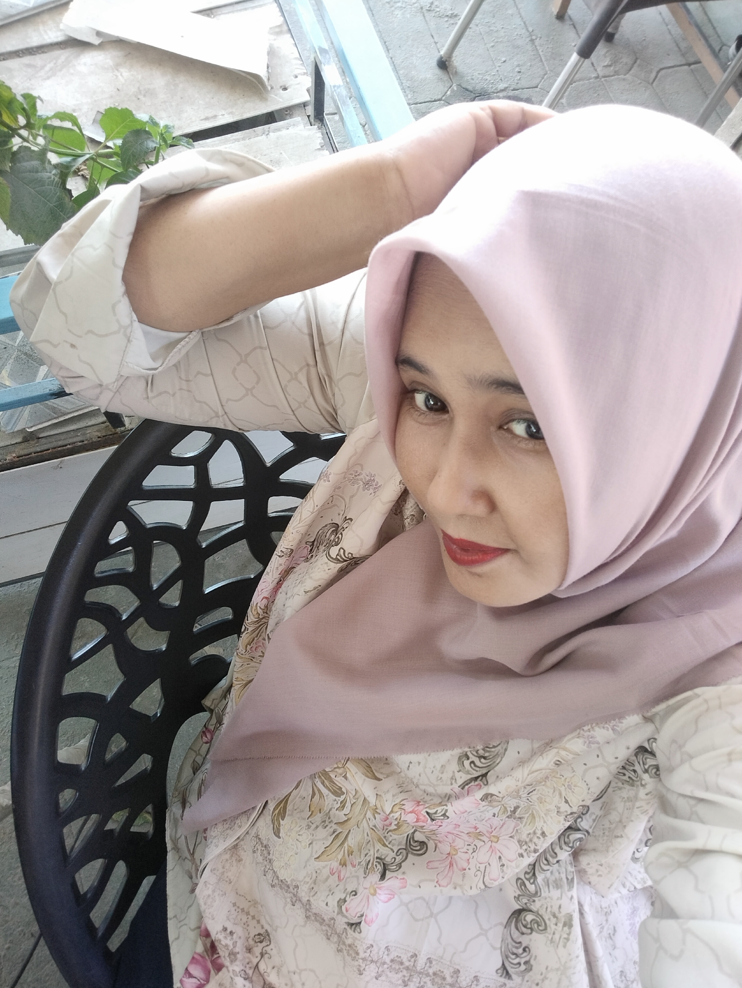 A woman wearing a light pink hijab and a floral patterned top is seated in a black, ornate chair. She is resting her arm on the back of the chair, and looking slightly downward with red lipstick. The background includes a plant with green leaves, a window frame with blue trim, and a partially visible surface with a gray stone texture. Based on the environment, the location is Matang, Indonesia.