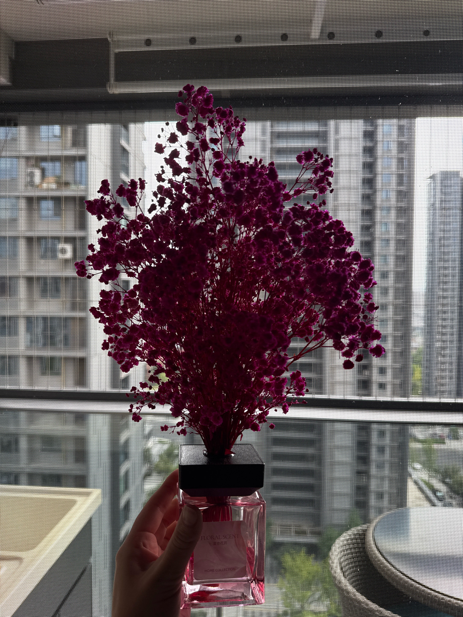 A hand is holding a pink glass bottle with a black lid, topped with a bouquet of dried purple flowers. The bottle has a label with Chinese text and the words "FLORAL SCENT" and "HOME COLLECTION." The scene is viewed through a window screen, with a blurred cityscape of tall buildings in the background. The environment appears to be an apartment or office building in Yiwu, China, given the characteristic architecture of modern Chinese cities and the presence of a window screen. A portion of a light-colored sink or countertop is visible on the left, and a woven outdoor chair and table set are partially visible on the right.