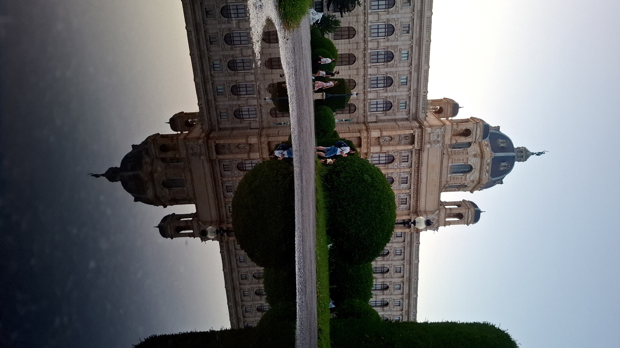 The image shows the Kunsthistorisches Museum in Vienna, Austria, as reflected in a puddle. The grand, ornate facade of the museum, with its dome and towers, is visible in the reflection, along with the sky and surrounding greenery. In the upper part of the image, the actual building is partially visible, with people walking on a path in front of it. The overall impression is one of architectural grandeur and the serene beauty of a reflection in water.