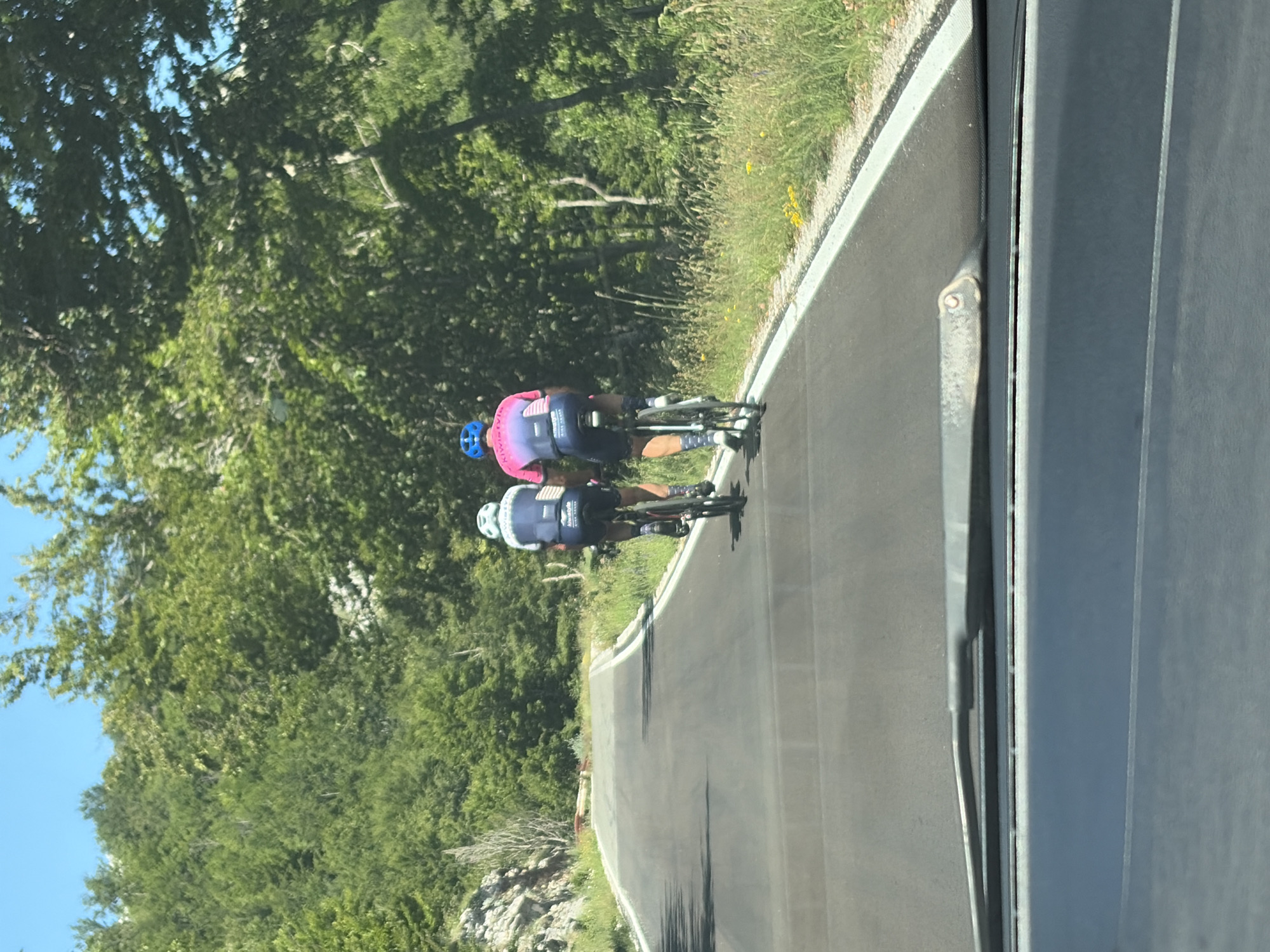 This image depicts a scene from inside a vehicle looking forward, capturing two cyclists on an asphalt road in Mali Bostur, Montenegro. The road, which appears to be on an incline, has a white solid line marking its right edge.
Two male cyclists are visible from behind, riding in the same direction as the camera vehicle. The cyclist on the left wears a blue and white jersey with "KIWISTYLE" and "RIDE SAFE" text, black cycling shorts, white socks, and a light blue helmet. The cyclist on the right wears a pink and blue gradient jersey with "KIWISTYLE" text, black cycling shorts, white socks, and a blue helmet. Both are riding dark-colored road bicycles.
The environment is characterized by dense, lush green deciduous forest covering hills on both sides of the road, extending under a clear blue sky. Rocky terrain is visible on the left side of the road. The roadside features green grass, weeds, and some yellow wildflowers. A white wooden fence post and a section of a red and white barrier are visible further along the road on the right and left, respectively. The bottom portion of the image shows the dashboard and a windshield wiper of the vehicle from which the photograph was taken. The scene is brightly lit by natural sunlight, casting shadows.