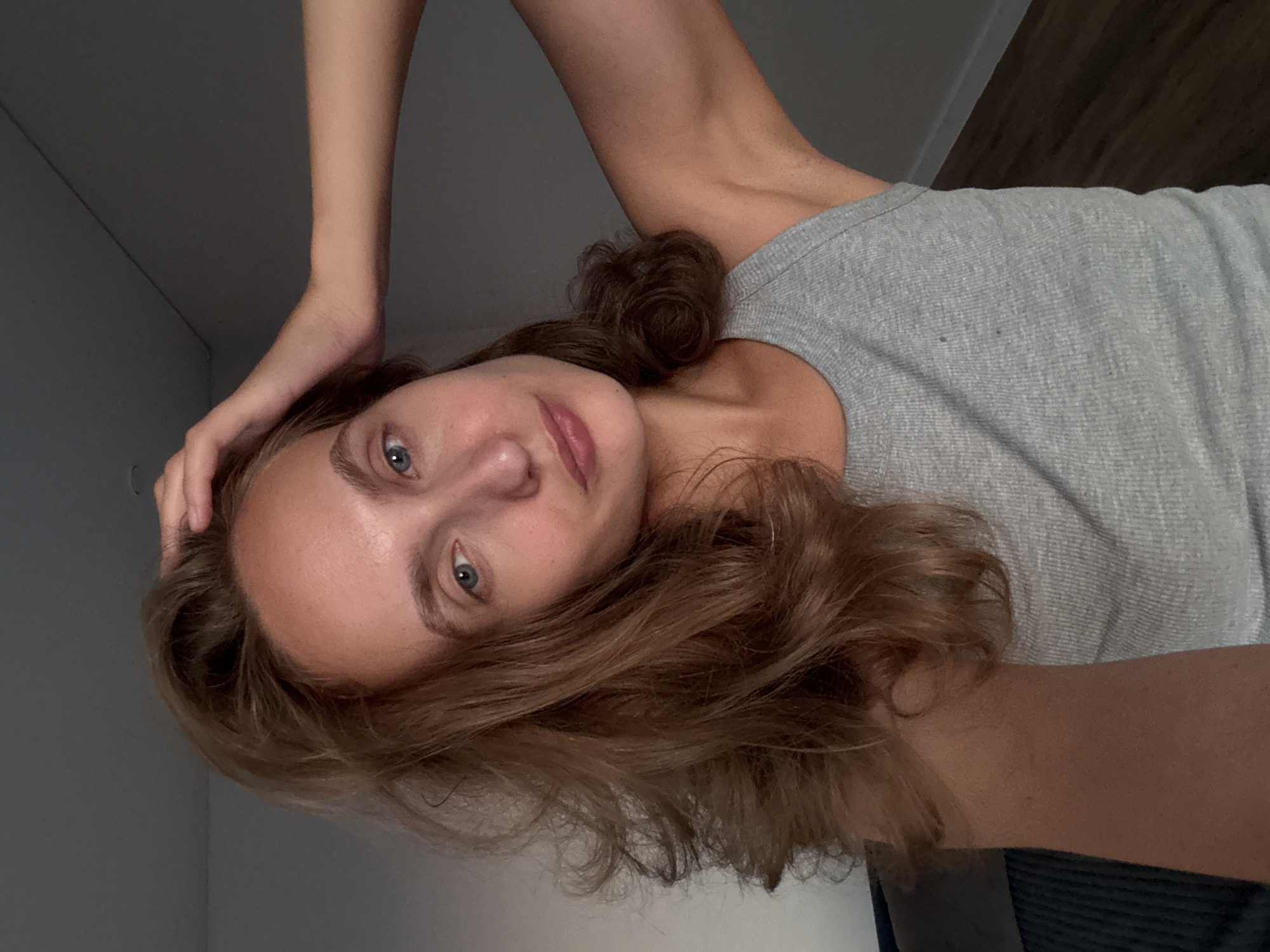 A young woman with medium-length, wavy, reddish-brown hair is shown from the chest up, looking towards the camera. Her right hand is raised, touching her hair at the crown of her head. She is wearing a grey, ribbed tank top. The background is a plain, muted grey wall with a dark wooden floor visible in the lower right corner. The scene appears to be indoors, in a dimly lit room. There are no discernible landmarks or indicators of the specific location being Подшивалово, Russia.
