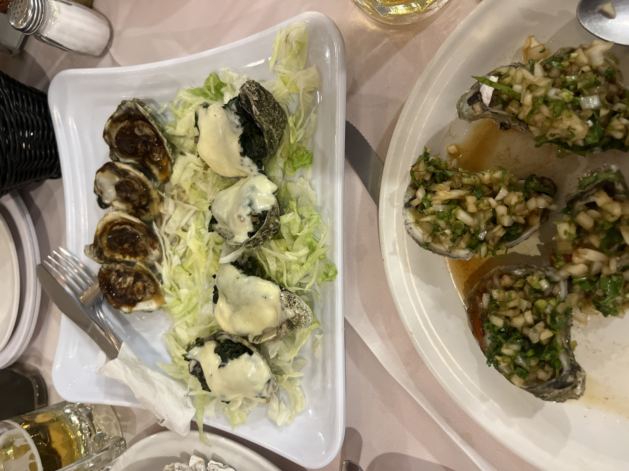 The image shows two plates of oysters, likely taken in a restaurant. One plate contains six oysters, three topped with a white sauce, and three that appear to be baked. They are arranged on a bed of shredded lettuce. Silverware rests near the oysters. The second plate features oysters covered with a green salsa. The scene includes glimpses of other dining implements, and a salt shaker. While it is difficult to determine exact location with given information, this could be in San Bernardino Tlaxcalancingo, Mexico, based on the instructions.