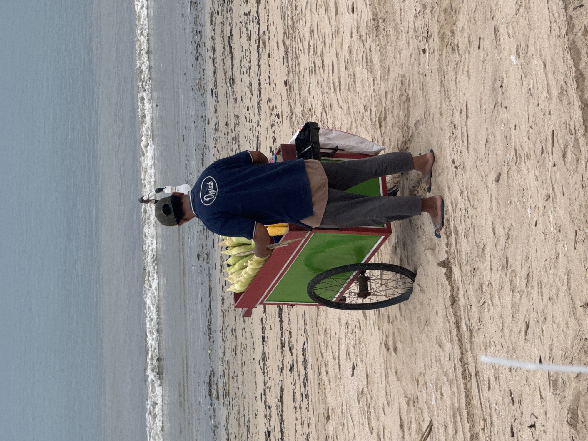 A person stands on a sandy beach near the ocean at Kuta Beach in Kuta, Indonesia. The person is wearing a dark blue shirt with white writing on the back, grey pants, a hat, and blue flip-flops. They are standing next to a green and red cart that is filled with ears of corn. The ocean is to the left of the person, and the sand is to the right.