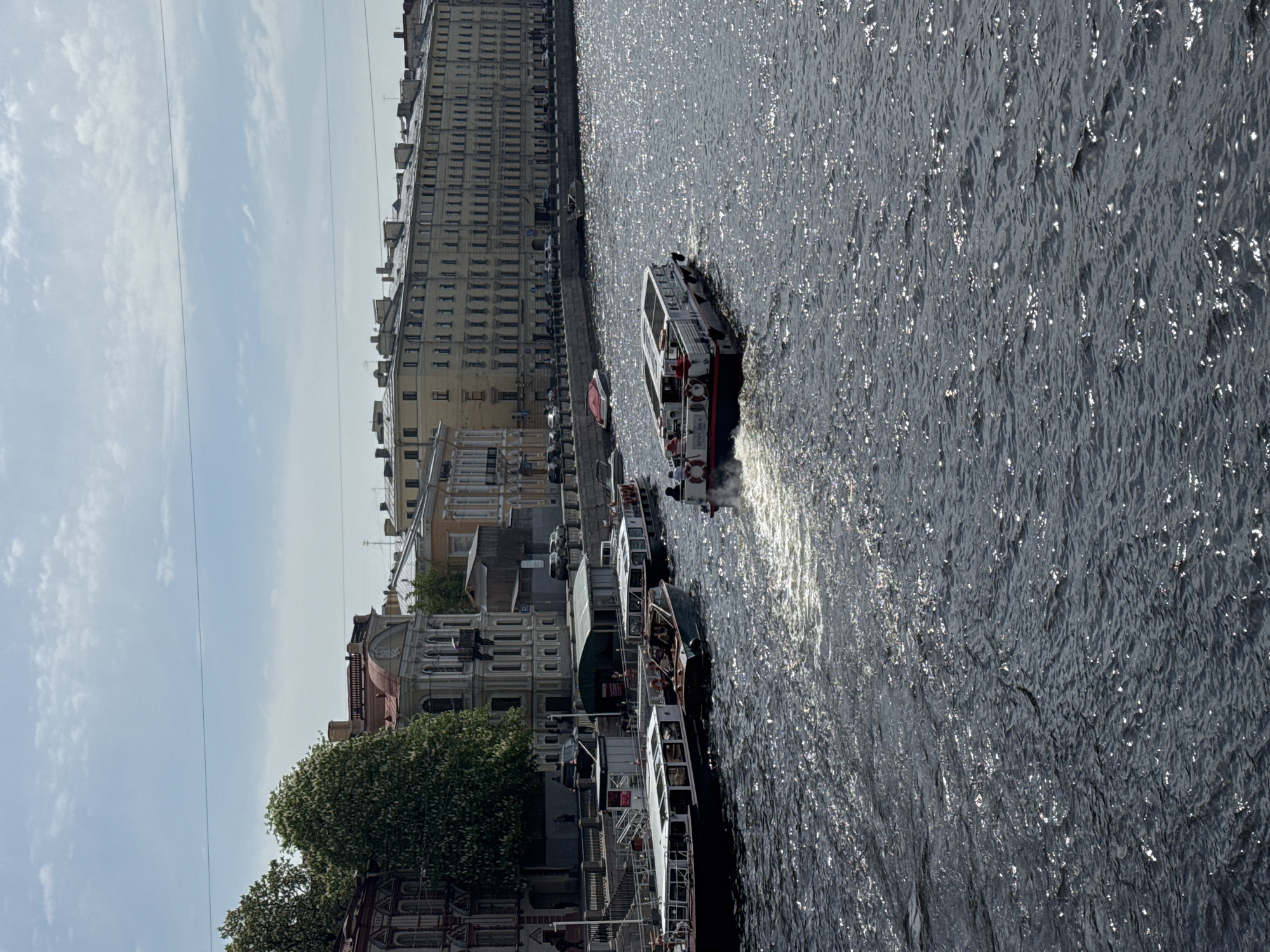 The image depicts a waterscape in Saint Petersburg, Russia. The view includes a row of historic buildings lining a riverbank, with several tour boats docked along the embankment and one moving through the water. The water surface reflects sunlight, creating a shimmering effect. The boats are occupied by people. The overall atmosphere is one of a busy waterfront scene in a historic urban setting.