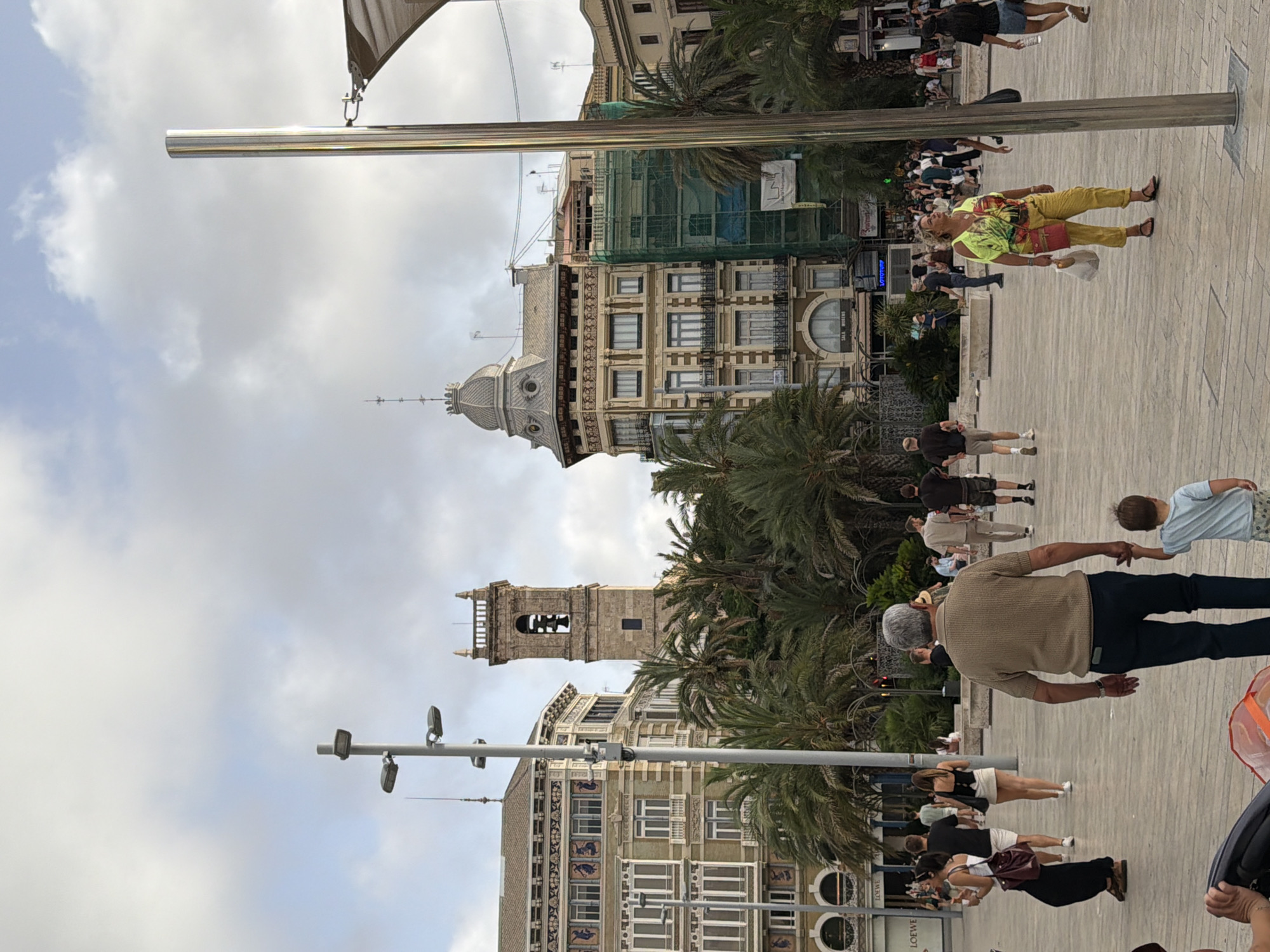 The image depicts an outdoor public square in a European city, likely during daytime with overcast skies. In the background, several buildings are visible, including a prominent bell tower with an arched opening containing a bell, and a building with a dome-like structure and intricate architectural details. Another building is partially covered in green scaffolding. Palm trees are scattered throughout the square, suggesting a warm climate.
The foreground and midground are populated by various individuals. In the immediate foreground, an adult male wearing a brown sweater and dark pants is walking while holding the hand of a young child in a light blue shirt and patterned shorts. To the left and mid-distance, a woman in a white top and black skirt walks away from the camera, carrying a dark red purse. Behind her, a group of people are walking in different directions, some casually, others appearing to be tourists. Further to the right, a woman in a brightly colored yellow outfit and a multi-colored top is looking upwards, holding a white shopping bag and a red purse. Several other individuals are scattered throughout the square, some sitting on benches, others walking. The ground is paved with light-colored square tiles. Lampposts and a tall metal pole are also present in the scene. The overall atmosphere appears to be one of leisurely public activity.
