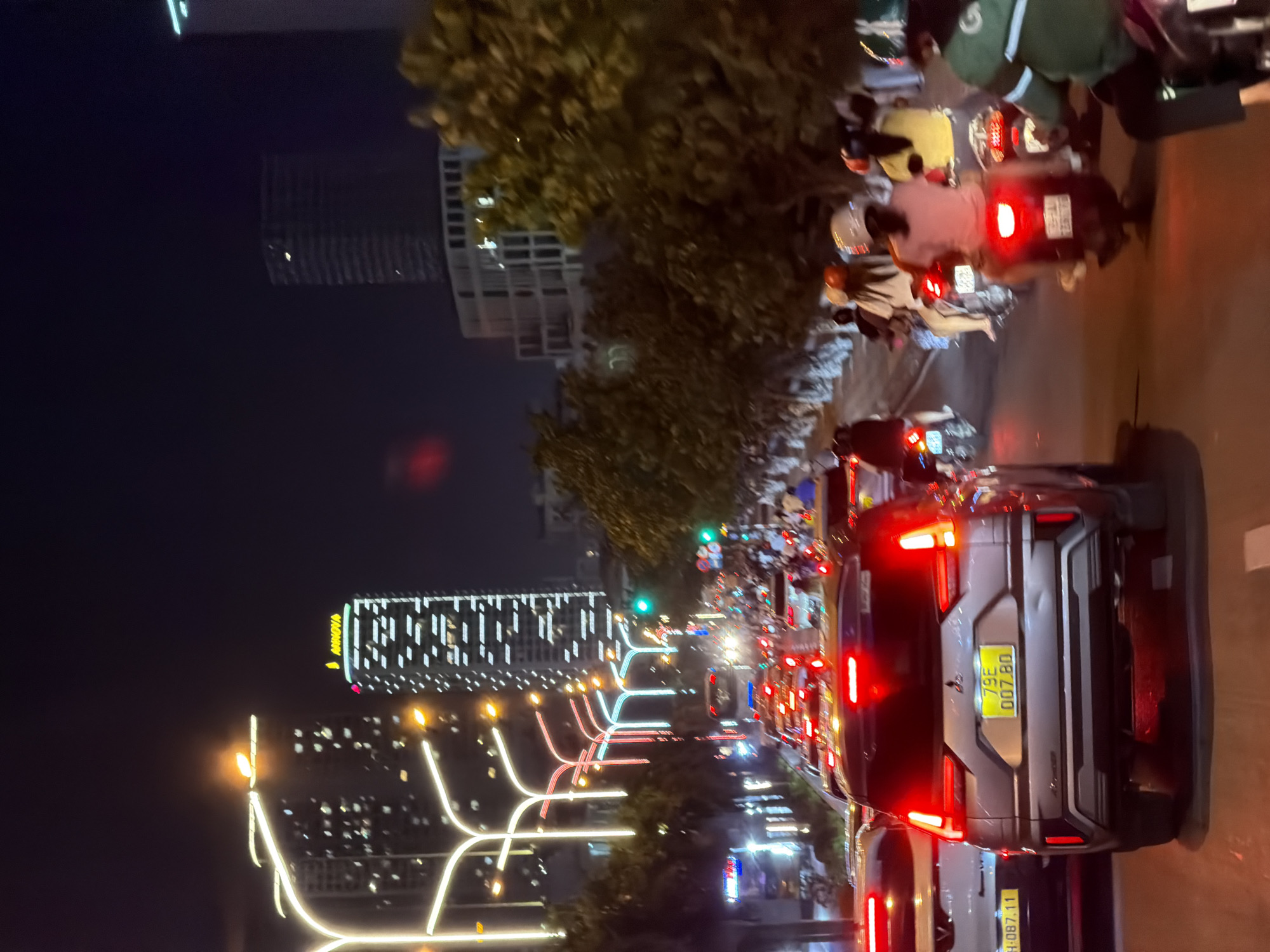 The image depicts a busy urban street scene at night in Bac Nha Trang Ward, Nha Trang, Vietnam. The perspective is from a vehicle stopped in traffic, looking forward into a dense flow of cars and motorcycles.
In the immediate foreground, the rear of a dark grey Mitsubishi Xpander SUV is central, its red tail lights brightly illuminated. The vehicle displays a yellow Vietnamese license plate, "79E 007.80." A minor dent is visible on the lower left portion of its tailgate. To the left of the SUV, the rear section of a white vehicle is partially visible, bearing the license plate "79A H-087.11."
The street ahead is congested with a multitude of vehicles, characterized by a concentration of red tail lights extending into the distance, along with scattered white headlights. On the right side of the frame, a distinct lane of motorcycles moves parallel to the cars. Several motorcycle riders are visible, many wearing helmets. The closest rider on the right wears a green jacket, potentially indicating a delivery service.
The thoroughfare is brightly lit by distinctive, stylized streetlights with curved white-yellow illuminated fixtures. Additional decorative lighting in white, red, and blue is present on poles along the street. Mature trees line both sides of the road, partially obscuring structures.
In the background, several modern high-rise buildings rise into the dark night sky. A prominent building in the mid-ground features an illuminated yellow "ANNOVA" sign at its top. Other buildings display numerous lit windows, creating patterns. A green traffic light is visible on the right side of the street further down, indicating active traffic flow. The scene conveys a bustling urban environment illuminated by artificial light.