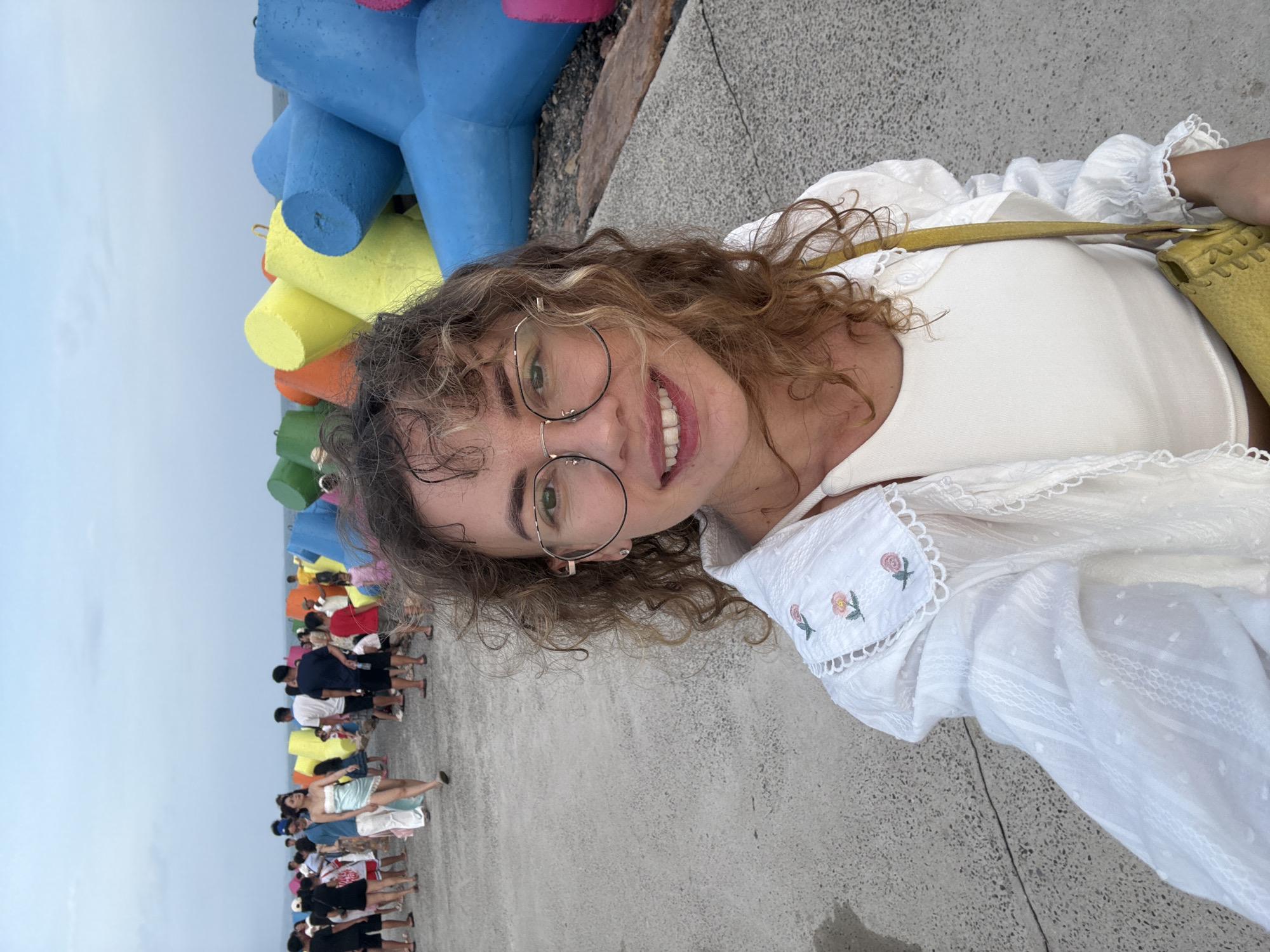 In a selfie taken outdoors, a woman with curly brown hair and glasses smiles at the camera. She is wearing a white top and a white open-front shirt with floral embroidery on the collar. A yellow purse is slung across her shoulder. Behind her, a paved walkway stretches into the distance, lined with colorful tetrapods that serve as breakwaters. A crowd of people walks along the walkway, some in casual attire, suggesting a public gathering or recreational area. The sky is overcast, and the sea is visible in the background. The location is identified as a public area with breakwaters in an unknown city in Vietnam.