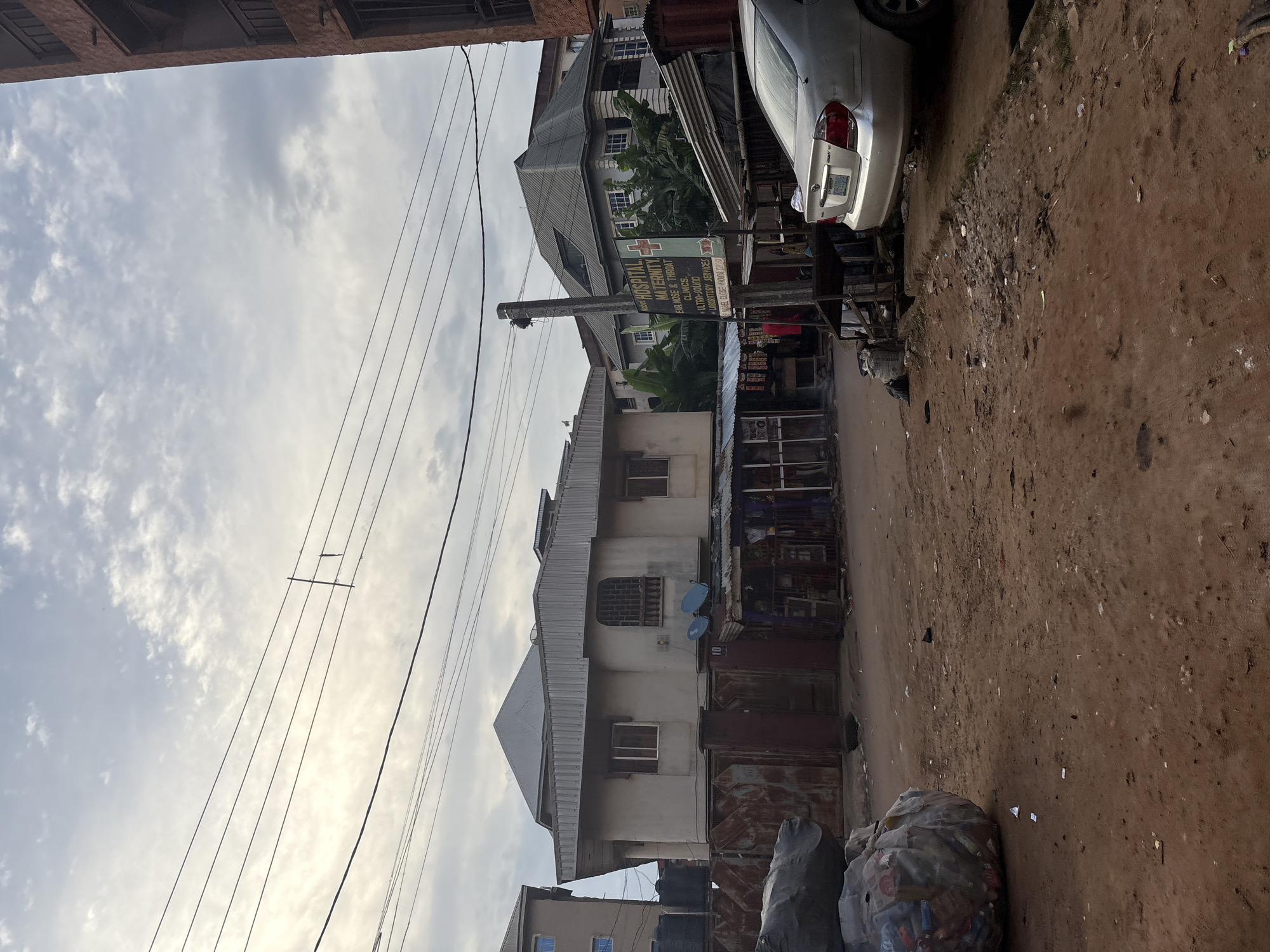 The image depicts a street scene in Onitsha, Nigeria, under an overcast sky. A large, multi-story building with a grey, pitched roof dominates the center-left of the frame. It has several windows and a white facade, with a rusty metal fence in front. Adjacent to it, to the left, is another building, taller and lighter in color, with visible water tanks. To the right of the central building, a lighter colored building with a grey roof is visible, partially obscured by vegetation.
In the foreground, a dirt road shows signs of disrepair and scattered debris. On the far left, a large, compressed bale of what appears to be plastic waste is a prominent feature. A silver sedan is parked on the right side of the road, facing away from the camera.
A signpost with red lettering on a white background, mounted on a pole, is visible to the right of the central building. It appears to advertise medical services, with text including "HOSPITAL", "MATERNITY", "EAR NOSE & THROAT CLINICS", and "ULTRASONIC LABORATORY SERVICES". Small market stalls with corrugated metal roofs are set up along the roadside in front of the buildings. Overhead, power lines crisscross the sky. No people are clearly visible in the scene.