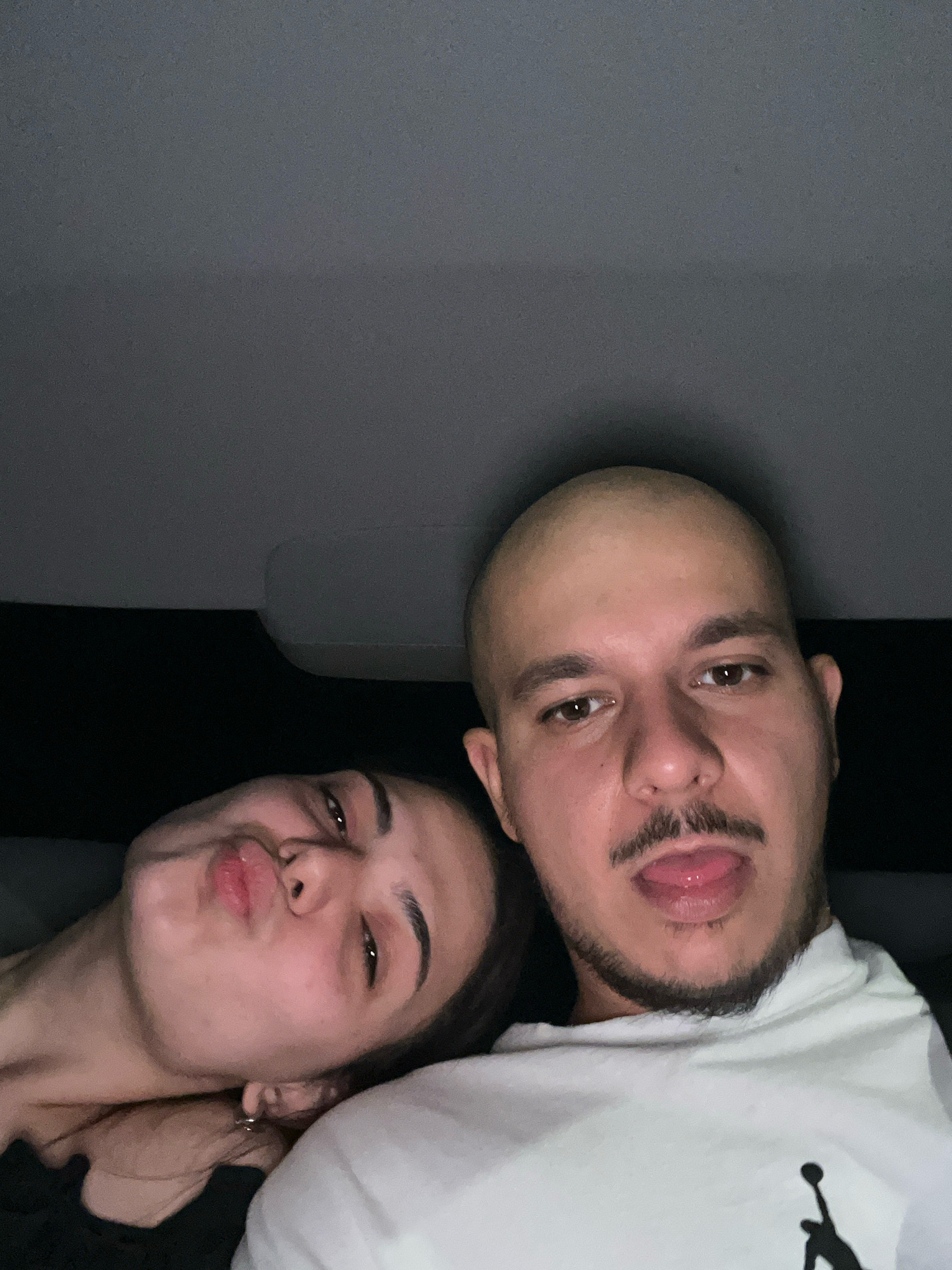 In a car, a bald man with a mustache and goatee has his tongue sticking out, looking at the camera. Next to him, a woman with dark hair leans her head on his shoulder, puckering her lips and looking away from the camera. They appear to be in the backseat of the vehicle, with the ceiling of the car visible above them.