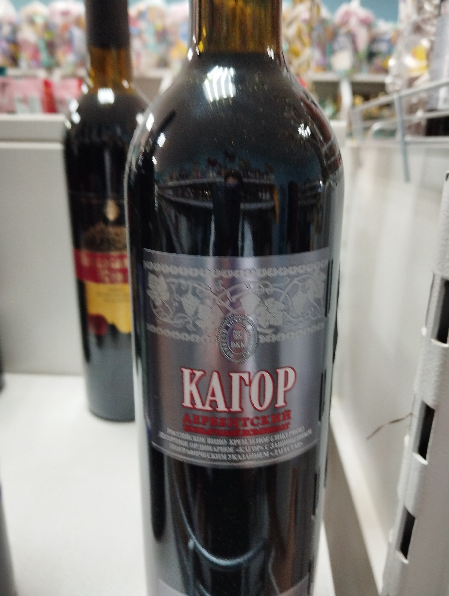 Here's a detailed description of the image:
**Scene:** The image shows two bottles of wine, with the focus on the one in the foreground. The background is a blurred retail environment, possibly a store.
**Objects:**
* **Wine Bottles:** Two bottles are visible. The front bottle has a silver label with red lettering that reads "КАГОР" (Kagor) in Cyrillic script, followed by "АРБАТСКИЙ" (Arbatsky). The label also contains finer print and decorative elements. The bottle in the background has a red and yellow label.
* **Shelf:** The bottles appear to be sitting on a white shelf.
* **Retail Environment:** Behind the bottles are blurred shapes suggesting shelves and products, indicative of a retail setting.
**Environment:**
* **Lighting:** The scene is brightly lit. The reflections on the front bottle suggest overhead lighting.
* **Location Clues:** The label on the wine bottle indicates it is "Kagor Arbat", which is a type of fortified wine. The Cyrillic script and the name "Arbatsky" suggest that the location might be in a Russian-speaking region.
**Implied Information:**
* **Purpose:** The bottles are likely for sale, as they are displayed in a retail setting.
The product appears to be "Kagor Arbatsky" wine, commonly associated with Eastern European winemaking traditions. The overall image suggests a consumer setting, such as a wine shop or grocery store.