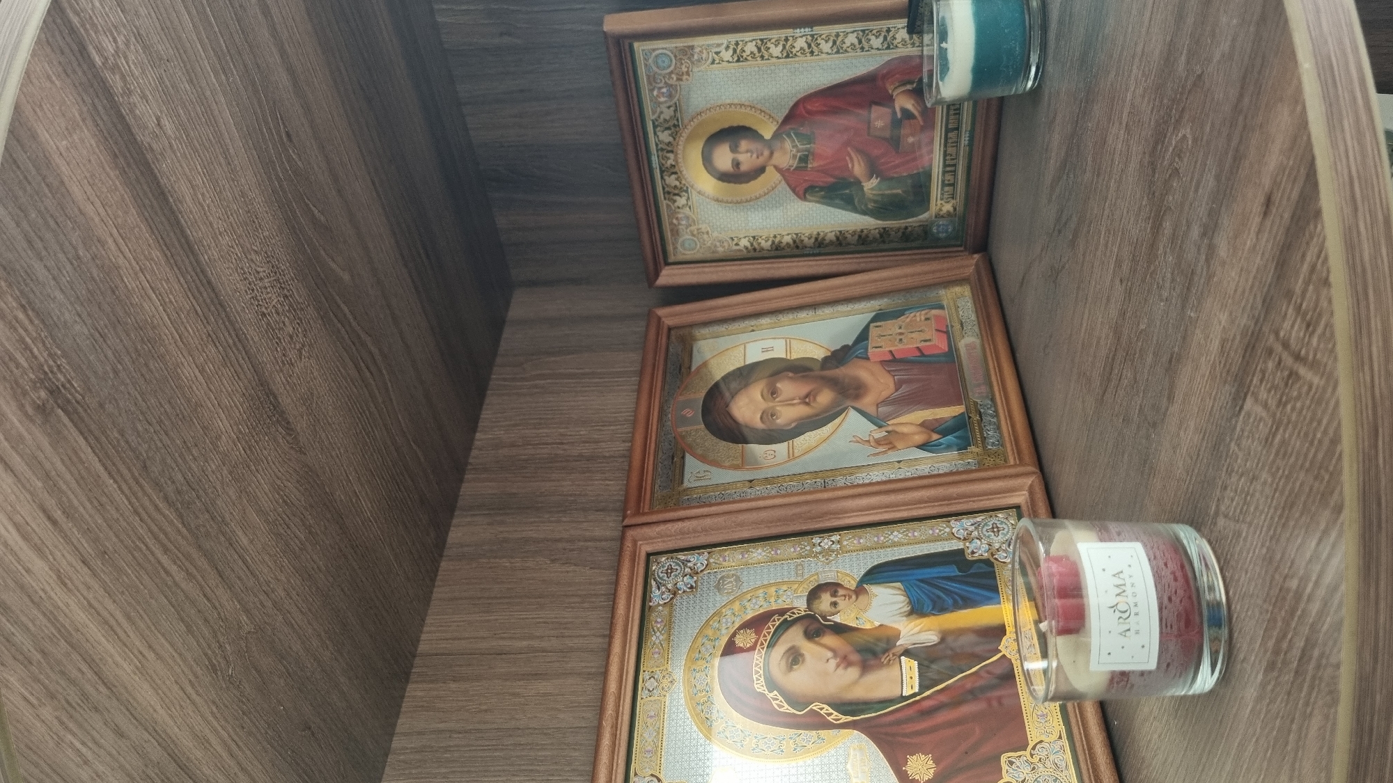 The image shows three framed religious icons and a candle on a wooden shelf. The icons depict the Virgin Mary with the infant Jesus, Jesus Christ, and another male figure, possibly a saint. All figures are rendered in a traditional iconographic style. The shelf is made of a wood-like material with a visible grain pattern. A glass candle with a red and white wax filling and a label that reads "AROMA HARMONY" is placed in the foreground to the left. To the right, a small blue and white candle is visible. The background consists of a darker wooden surface, suggesting this is inside a cupboard or on a bookshelf. The overall setting appears to be domestic. The location is stated to be Верзилово, Russia.