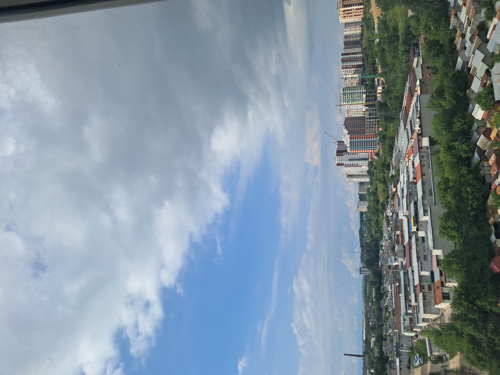 This is a high-angle, wide shot of a cityscape with a mix of residential buildings and industrial elements under a cloudy sky. In the foreground, a dense cluster of low-rise, uniform buildings with predominantly grey walls and varied roofing materials like rusted metal and red tiles are visible. These buildings appear to be residential units or garages. Further into the midground, more of these low-rise structures are present, interspersed with green trees. In the background, a row of taller, modern apartment buildings with colorful facades and multiple stories dominates the horizon, some of which are under construction, indicated by the presence of cranes. To the left, a tall, narrow industrial smokestack is visible, hinting at industrial activity in the vicinity. The sky is a mix of bright blue with scattered white clouds on the left, transitioning to darker, heavier grey clouds on the right. The overall scene depicts a developing urban area with both older and newer construction.