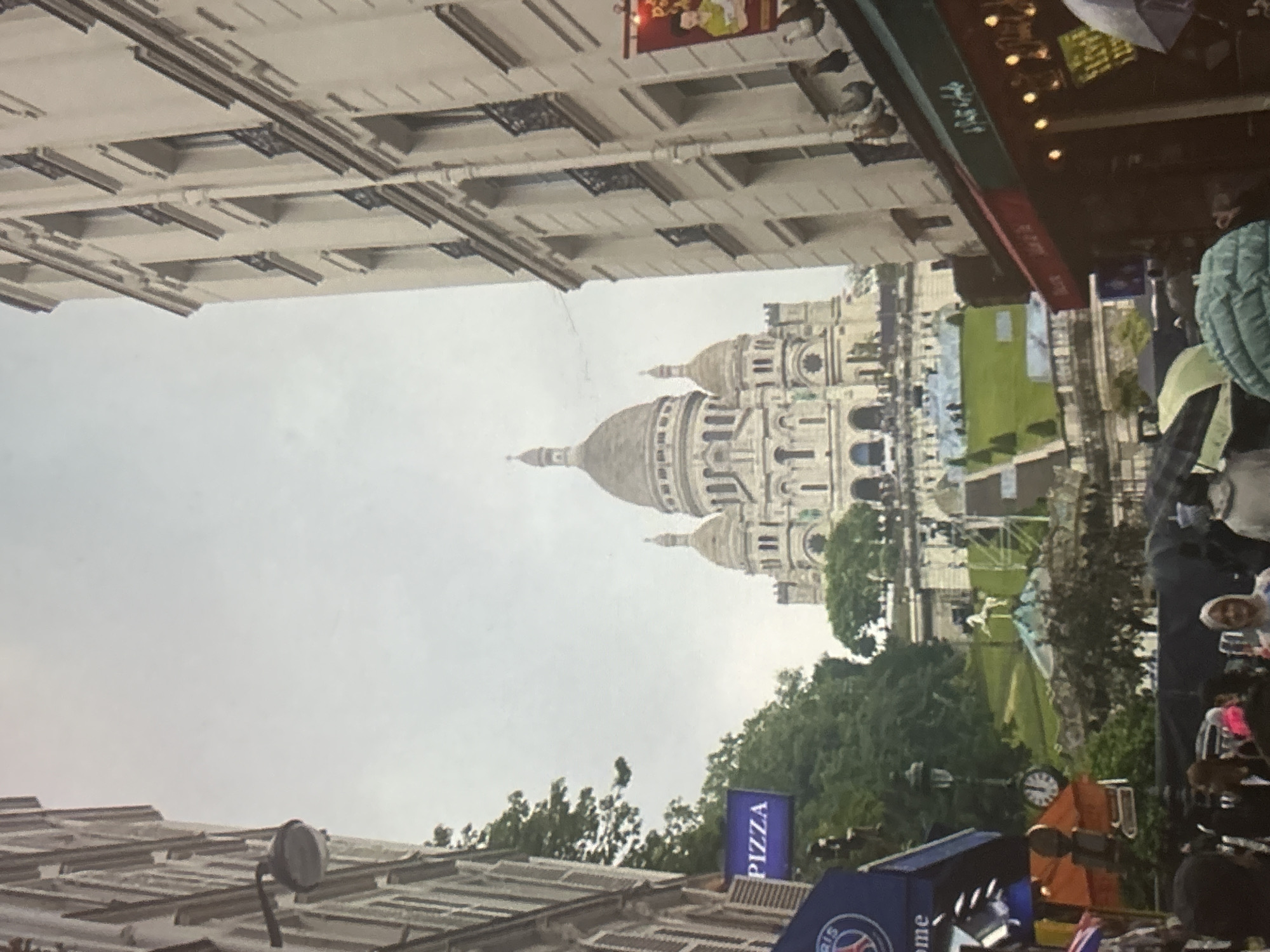 The image depicts a street view in front of the Sacré-Cœur Basilica in Paris, France. The basilica, with its iconic domes and white facade, dominates the background. In the foreground, a street is lined with buildings on both sides. On the left, a building displays a blue sign that reads "PIZZA". A black lamp post with a clock is also visible. People are walking on the street, some holding umbrellas, suggesting it might be a cloudy or rainy day. On the right side of the street, there is a building with awnings, possibly housing shops or restaurants. The overall scene captures a bustling urban environment with a prominent historical landmark.
