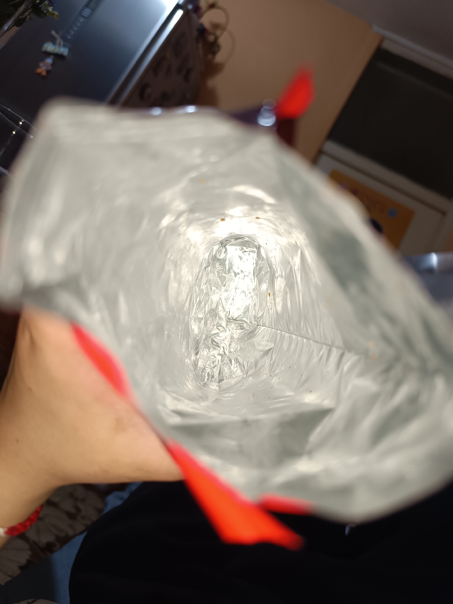 The image shows the inside of an empty bag of potato chips. The bag is metallic silver on the inside. There are a few crumbs visible at the bottom of the bag. Red closure clips are visible at the corners of the bag. An arm and wrist wearing a red beaded bracelet is visible. The background is out of focus, but shows a kitchen counter and cabinets. There is not enough information to determine the specific location in Pleven, Bulgaria.