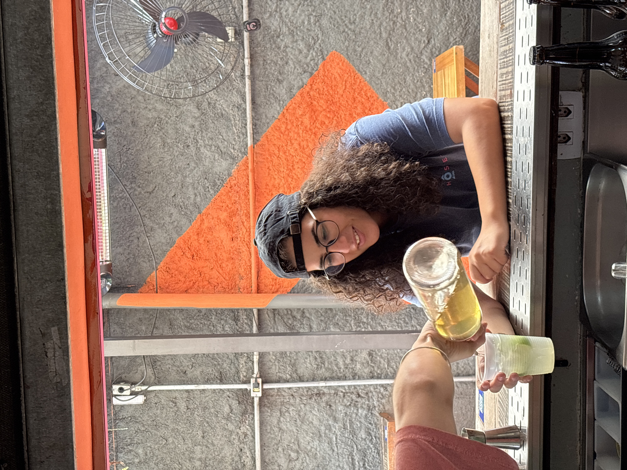 A low-angle indoor shot shows a person with curly hair and glasses wearing a black baseball cap, smiling while pouring liquid from a clear bottle into a clear plastic cup held by another person, likely a customer. Both are standing at what appears to be a bar. In the background are a concrete ceiling, an orange triangle, a metal fan, and electrical fixtures. The photo was taken in the city of São Paulo, Brazil.