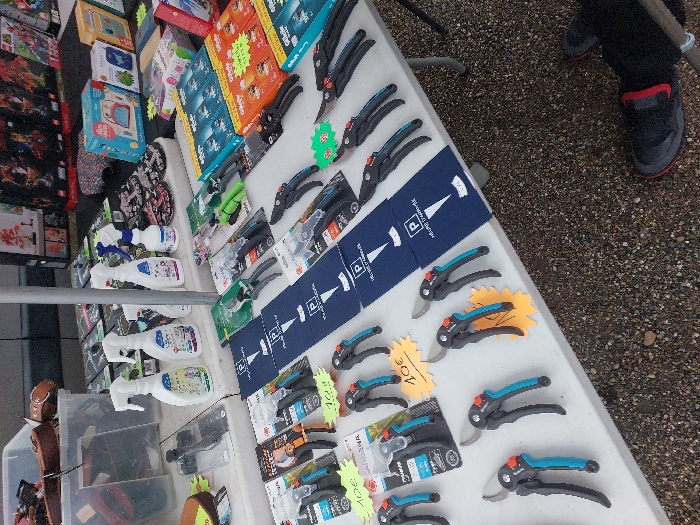 The scene depicts a market stall in Eckbolsheim, France, exhibiting various merchandise. The primary subject is a white table laden with goods. In the foreground and mid-ground, multiple pairs of black and blue/teal handled gardening secateurs/pruners are displayed, both packaged and unpackaged, with several bearing yellow price tags indicating "10€." A row of blue parking discs, each printed with a white "P" and "HEURE D'ARRIVEE," is laid out centrally. To the left, several white plastic spray bottles, labeled as household cleaning products, are visible.
Further back on the table, rows of packaged items, including boxes of Gillette razor blades and small boxes for children's items (e.g., "Goutte d'eau"), are arranged. In the background, additional merchandise is visible, including boxed sets resembling construction toys. A vertical metal pole, likely a support for a market canopy, is present on the table's left side. The ground beneath the table is composed of gravel or small stones. A person's right foot, encased in a dark shoe with a red accent, and part of their dark trouser leg, is visible in the lower right corner, positioned on the gravel next to the table.
