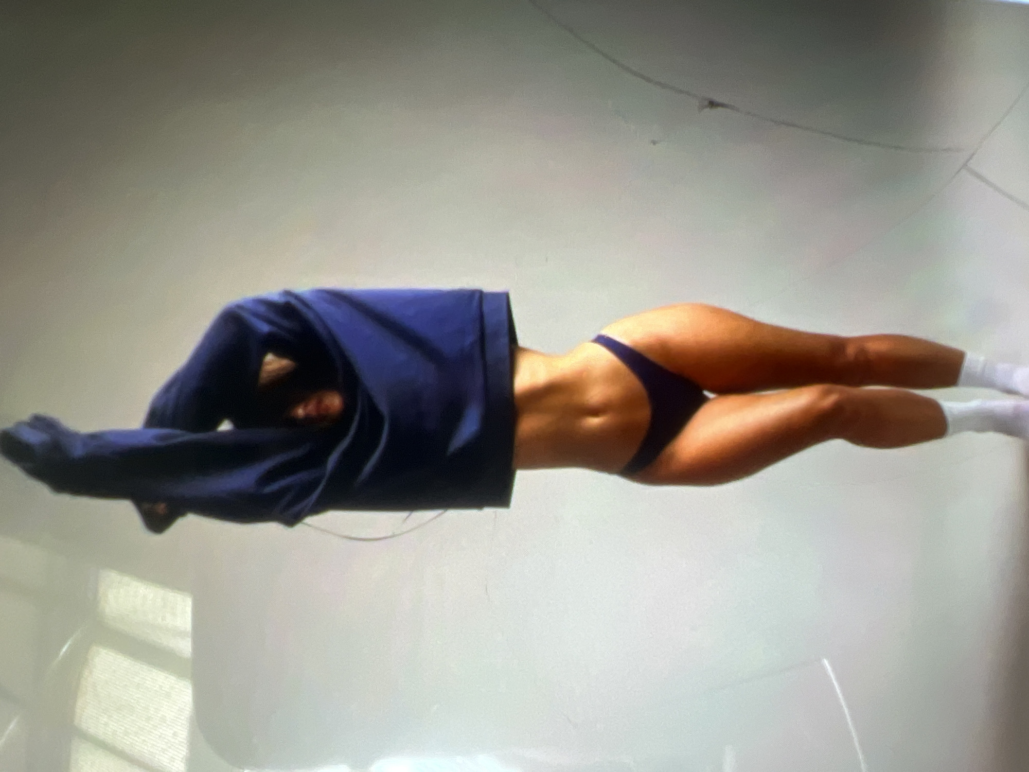 A person is standing with their arms raised, pulling a dark blue sweatshirt over their head. They are wearing dark bikini bottoms and white socks. The background is a plain, light-colored wall with some soft lighting. There is no indication of a specific location in Kara-Dıykan, Kyrgyzstan in the image.