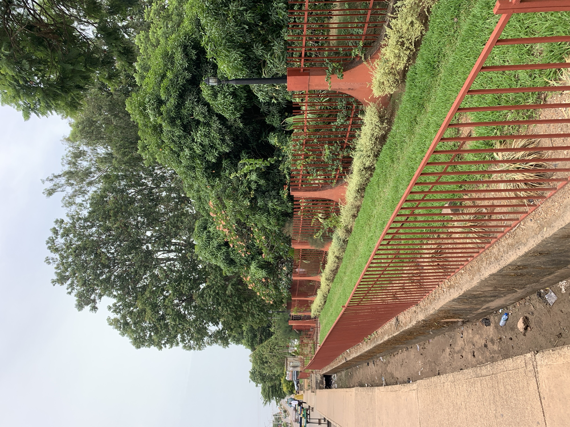 The image depicts a scene along a street in Barakin Lerawi, Nigeria. A red metal fence runs along a raised garden bed with green grass and various plants. A concrete walkway is present in the foreground with scattered trash. In the background, there are trees, buildings, and vehicles, and people can be seen further down the street. The sky is overcast.