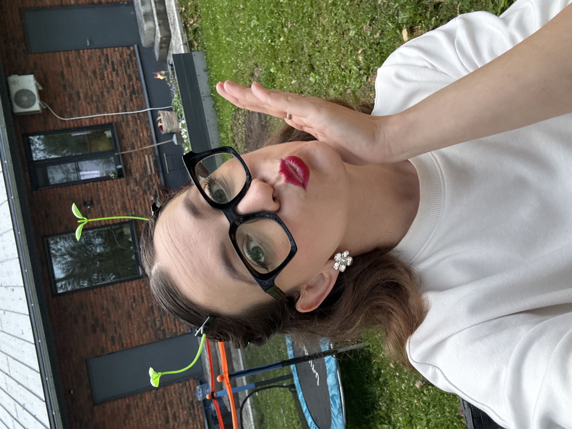 The image shows a young woman taking a selfie outdoors. She is wearing a white sweatshirt, black-rimmed glasses, and a flower-shaped earring. The woman has bright red lipstick on and is holding her hand up to her cheek. She has two green hair clips shaped like sprouting plants in her hair. In the background, there is a brick building with windows and an air conditioner unit, as well as a grassy area with some outdoor equipment, possibly a playground or trampoline. The scene appears to be set during the daytime, with natural light illuminating the area. The overall mood of the image seems playful and cheerful. There is no visible text in the image.