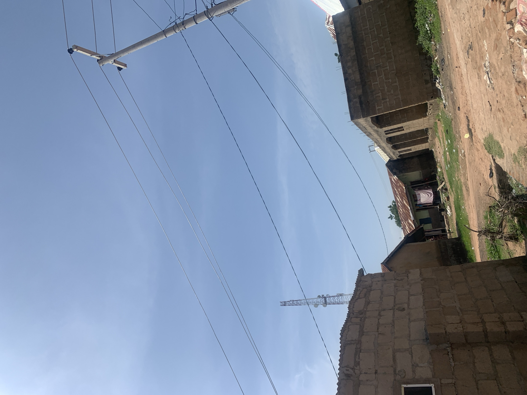 This is a photograph taken in Jauro Yinu, Nigeria, featuring a street scene under a clear blue sky. In the foreground, a rough, dirt road is visible, with debris scattered along its edges. To the left, a partially constructed building made of concrete blocks dominates the frame, with a corrugated metal roof. Further back, several other structures are present, some appearing to be residential homes with corrugated metal roofs and varying states of construction. A prominent feature of the image is a tall telecommunications tower in the distance, silhouetted against the sky. Electrical poles and wires are also visible, stretching across the frame and connecting to the buildings, suggesting the presence of electricity infrastructure in the area. The overall impression is one of a developing urban or semi-urban environment.