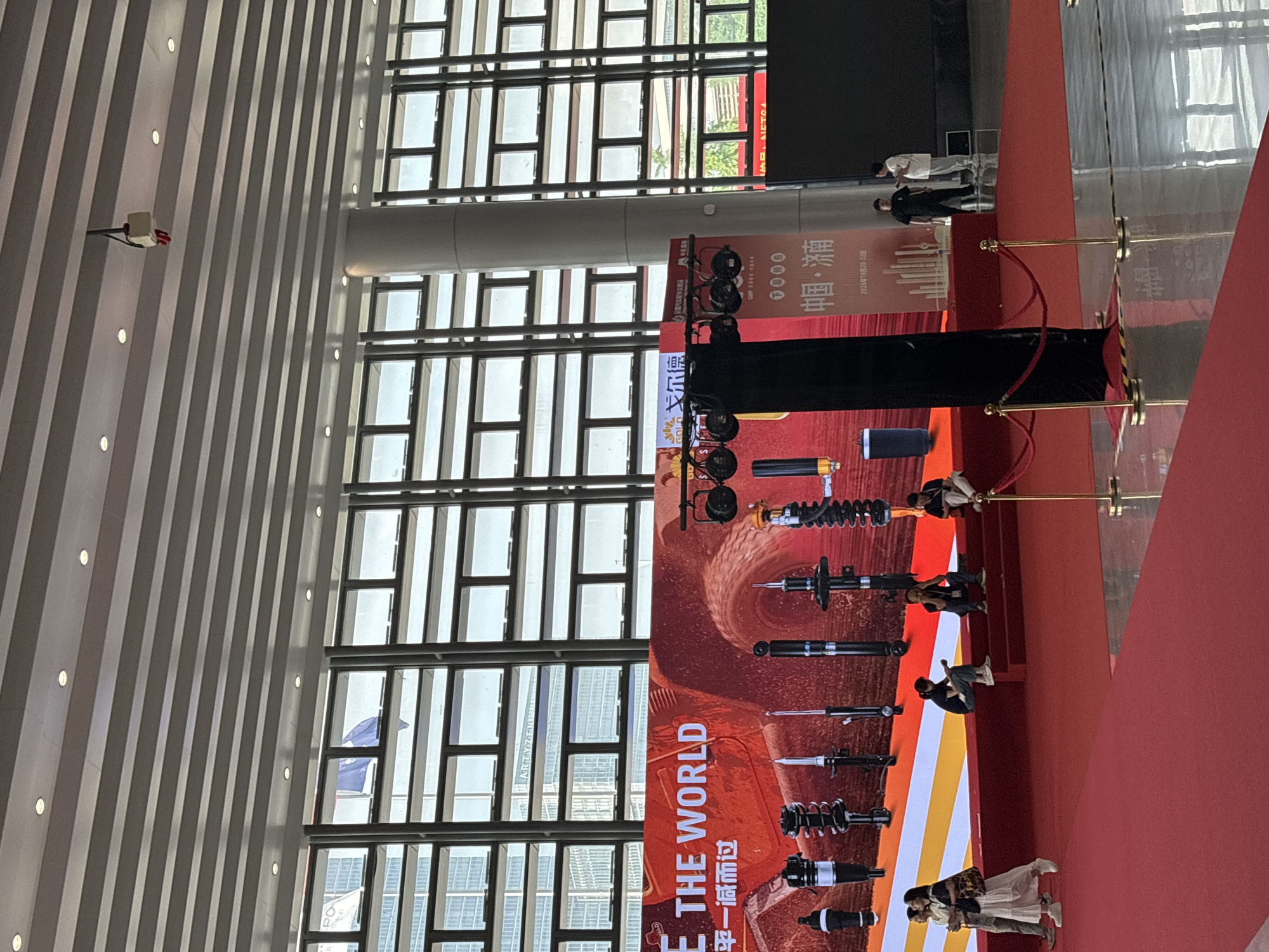 The image shows an advertisement display for "GOLD" shock absorbers, at an apparent trade show. The exhibition is set up in a spacious building, evident from the tall windows. The display includes the words "E THE WORLD" in large, bold letters, and the Chinese text “平一减而过”. Several shock absorber models are visible in the display.
Multiple individuals are present in the scene. Some people are walking by the display, appearing to be potential customers, while others are standing around or posing for photos near the shock absorber display. The red carpet leads up to the display.
The location is identified by the banner displaying "0000 帼·湳" and the date "2026年10月20-22日," suggesting an event in Liangjiang, China. Additional identifiers include "CAPY ERRORBAL" and "MONETCA."