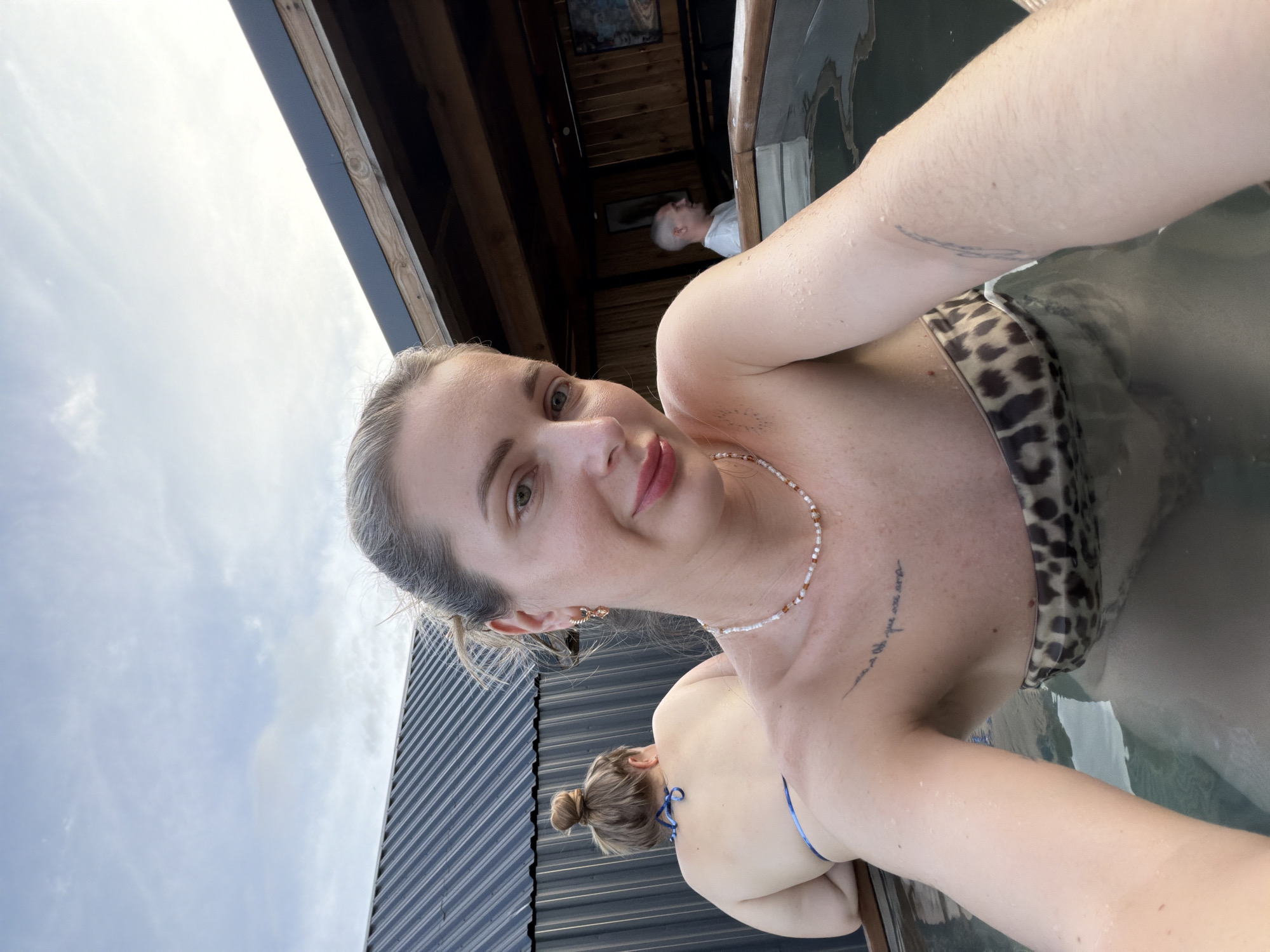 A selfie is taken by a blonde woman with a pearl necklace and cheetah-print bikini top, submerged in a hot spring. Behind her, another woman with her hair in a bun, wearing a blue bikini, leans on the edge of the pool. In the background, a man in a white t-shirt sits in the shade of a wooden structure. The scene appears to be an outdoor spa with a corrugated metal roof structure visible on the left and a wooden building with artwork on the right. The sky above is cloudy. The location is not identifiable as Семилуки, Russia.