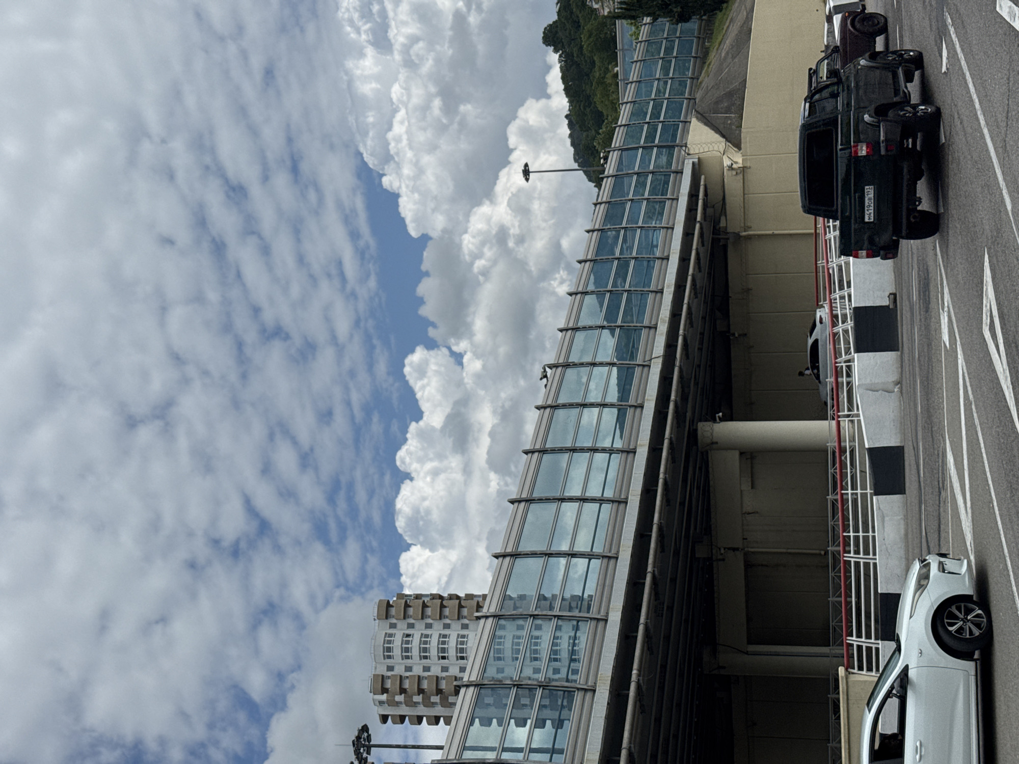 In Sochi, Russia, on a cloudy day, a black pickup truck with a license plate that appears to read "M4190B 193" drives on a paved road beneath an elevated structure. The elevated structure has a transparent, blue-tinted glass barrier along its side, reflecting the sky and the surrounding environment. To the left of the pickup truck, a white sedan is visible, also on the road. In the background, a tall, light-colored building with a grid-like facade stands out against the cloudy sky. Further in the distance, a lush green hillside is covered with trees, suggesting a natural landscape adjacent to the urban setting. The scene depicts a typical urban environment with modern infrastructure and vehicles.