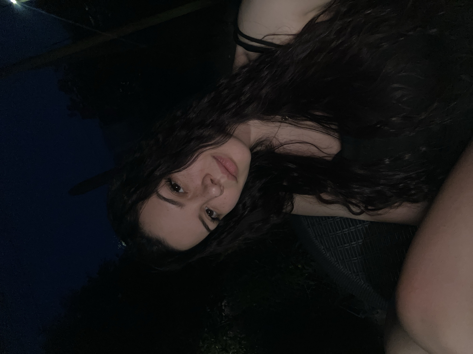 In Skala, Bulgaria, a young woman with long, dark, wavy hair takes a selfie outdoors at night. She is seated on a dark, woven chair, with the dimly lit background suggesting an outdoor setting with foliage and trees. She is wearing a black tank top. Her facial features, including her eyes, nose, lips, and freckles, are visible as she looks towards the camera. The overall lighting is low, with some artificial light source contributing to the illumination.