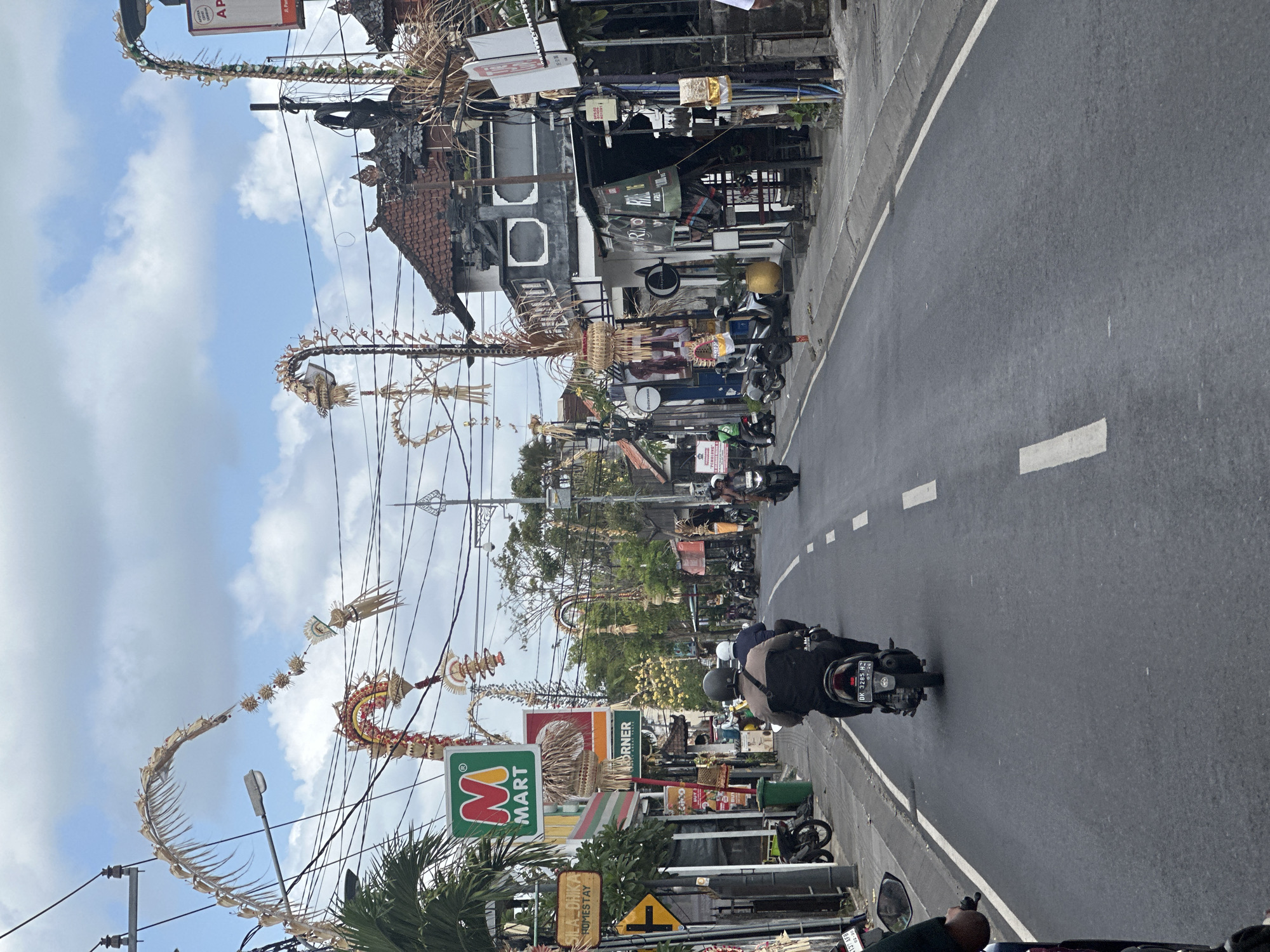 A street in North Kuta, Indonesia, is depicted, characterized by its asphalt roadway, sidewalks, and buildings lining both sides. Ornate, woven decorations, possibly made from palm leaves and resembling dragon-like forms, are strung across the street and attached to poles. Several motorbikes are present on the road, with some riders visible. One motorbike in the foreground has a license plate reading "DK 3285 HZ." Signs for "M MART" and "SILADIKA HOMESTAY" are visible on the left side of the street. The sky above is partly cloudy. The overall impression is of a bustling street scene with local cultural decorations.