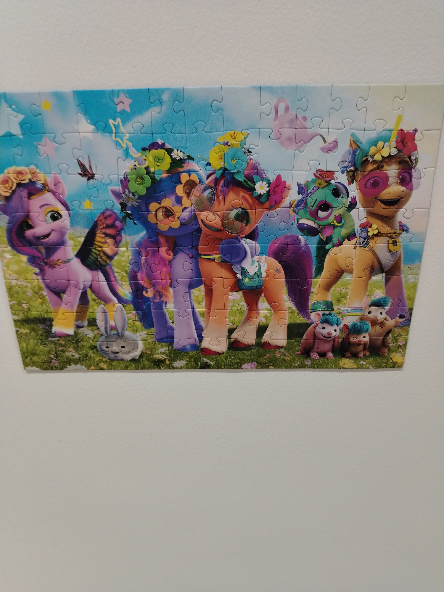 The image shows a puzzle depicting several My Little Pony characters and small animal companions standing in a grassy field with flowers under a blue sky with clouds and floating pink watering cans. From left to right, the characters include a purple pony with a floral crown and butterfly wings, a blue pony with a floral crown, an orange pony with a floral crown and sunglasses, and a yellow pony with a floral crown and sunglasses. In the foreground, there is a grey bunny and two small pink creatures with blue hair. The puzzle is hung on a plain white wall, and the lighting suggests an indoor environment, possibly a room. There is no indication of the location being in Dubai, United Arab Emirates, or any specific city.