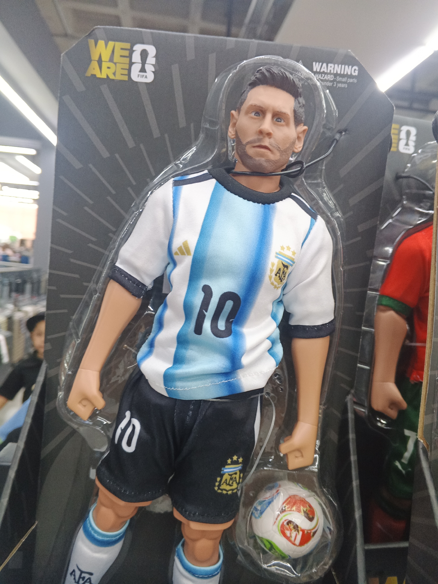 The image shows a Lionel Messi action figure in its packaging. The figure is wearing an Argentina national team jersey with the number 10. The packaging has the FIFA logo and a warning about small parts. Another action figure in a different soccer jersey is partially visible to the right. The image was likely taken indoors, possibly in a store, based on the shelves and lighting in the background. There is a person in the background to the left. There is no information in the image to suggest the location is Caracas, Venezuela.