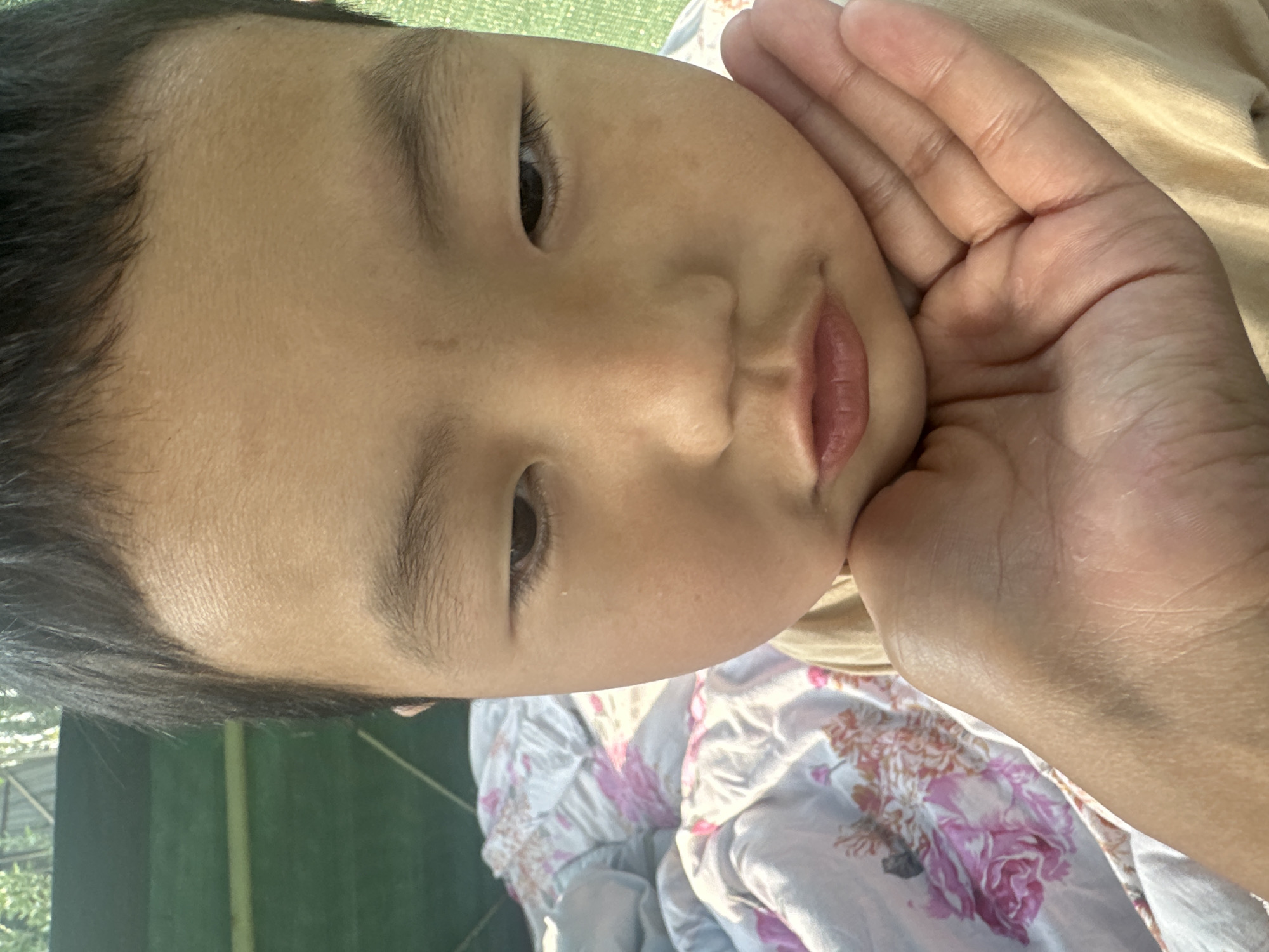 A close-up, overhead shot shows a young East Asian child with dark hair resting their chin on a person's palm. The child's eyes are looking down, and their lips are slightly pursed. The child is wearing a light tan shirt. The background consists of a blurred floral patterned comforter and a green surface, possibly a fence or wall. The scene is illuminated by natural light, casting shadows on the child's face. The location is specified as Taygarayev, Kyrgyzstan.