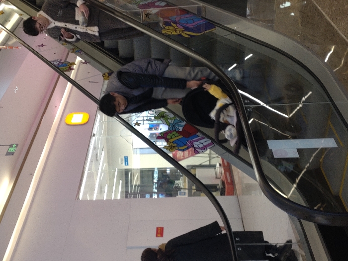 The scene depicts an upward-moving escalator within a commercial facility in Jiangjin, China. Three individuals are present on the escalator. In the mid-ground, a male subject, wearing a dark vest over a dark long-sleeved shirt and light trousers, is bent forward, attending to a dark-colored baby stroller positioned on the escalator step immediately beneath him. An infant, partially visible with light blankets and a yellow sleeve, occupies the stroller. On a step above, a second male subject, dressed in patterned light-colored loungewear, holds a mobile phone in his left hand and a rectangular object in his right, directing his gaze towards the phone screen. A third individual, seen partially from the back and left, wearing a dark coat, occupies a lower escalator step. The escalator features glass balustrades adorned with colorful gift-themed decals and a prominent white "1" sticker on a lower glass panel. In the background, a brightly lit retail store with a glass front and a distinctive orange oval logo is visible, displaying various merchandise. A red rectangular sign is affixed to the wall adjacent to the store entrance.