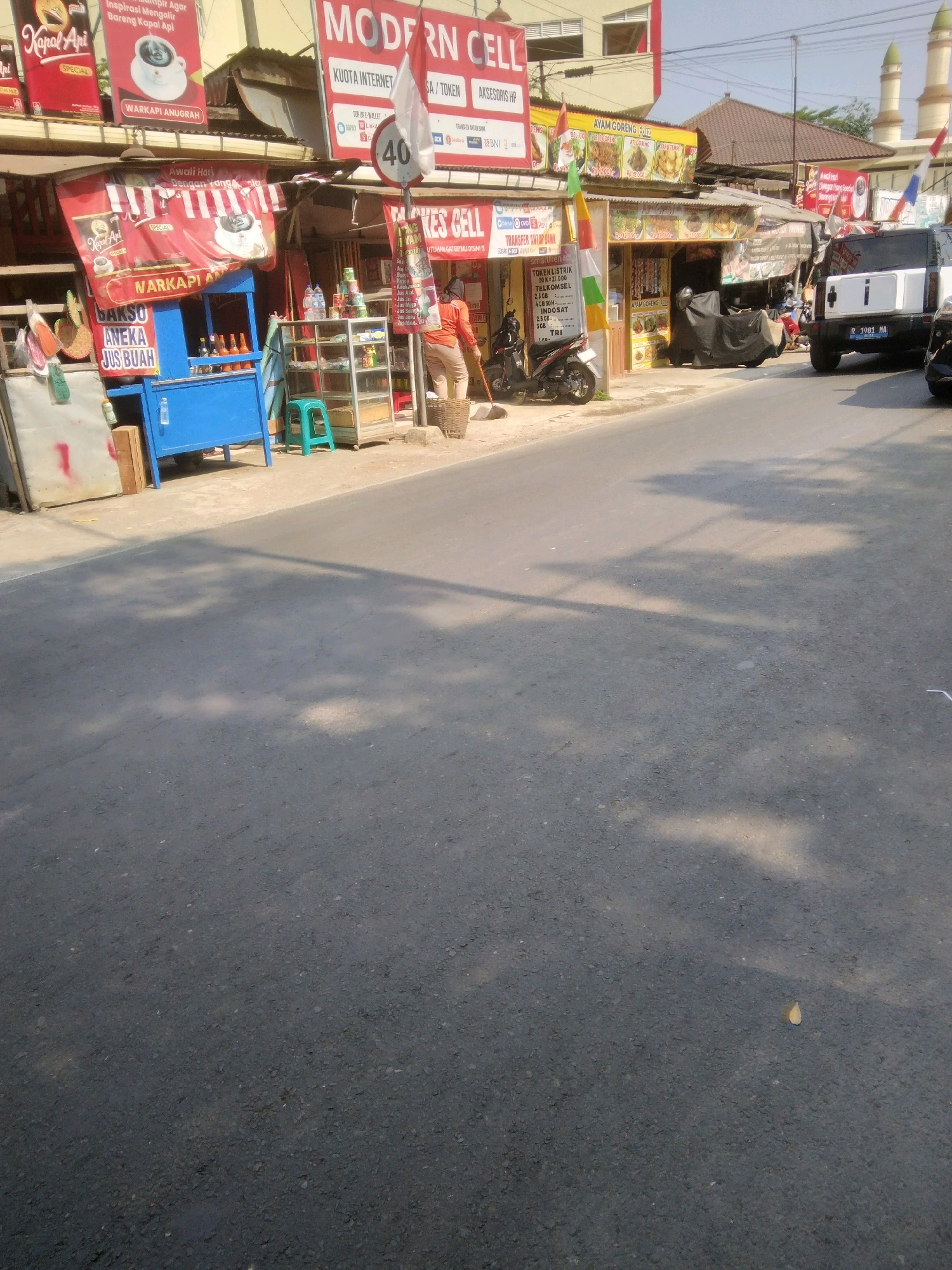 The image captures a street scene in Desa Pamijen, Kecamatan Sokaraja, Indonesia. The view is from street level, looking towards a row of small shops and stalls. In the foreground, the asphalt road is visible, with shadows of trees cast upon it.
To the left, a blue stall with a red banner is set up, advertising "NARIKAPI ANUGRAH" and "BAKSO ANEDA JUS BUAH". There are bottles and containers on display. A person in an orange shirt is seen in the middle of the frame, holding a broom and facing away from the camera, appearing to sweep the ground.
Above the stalls, advertisements are displayed. One large banner reads "MODERN CELL" with text underneath mentioning "KUOTA INTERNET", "TOKEN LISTRIK", and "AKSESORIS HP". Another banner advertises "AYAM GORENG". To the right, a white SUV with a license plate reading "R 1981 AA" is parked.
The environment suggests a busy, commercial area with multiple small businesses operating along the roadside. The architecture of the buildings appears simple, typical of many street-side establishments in Indonesia. The sky is clear and the lighting indicates it is daytime.