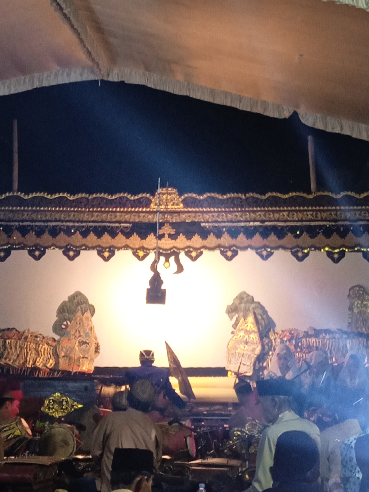 A traditional Wayang Kulit performance is underway, illuminated by a bright light source behind a translucent screen. Ornate decorations adorn the top of the screen, featuring gold and dark blue patterns. Figures, appearing to be puppets or actors, are visible on the screen, silhouetted against the light. In the foreground, a group of people are seated, some appearing to be musicians playing instruments such as drums, and others watching the performance. The setting appears to be outdoors under a tent-like canopy. The event is taking place in Mojotengah, Indonesia.