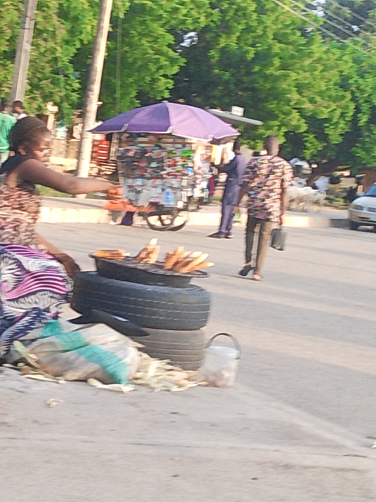 The media file depicts an outdoor scene, likely a street in Jalingo, Nigeria. The time of day appears to be late afternoon, with the sun casting long shadows and the light suggesting a warm, possibly humid, climate.
The foreground is dominated by a woman dressed in a patterned outfit, seated or kneeling beside a stack of tires. One tire serves as a base for a makeshift grill where several pieces of food, possibly corn on the cob, are being cooked. Beside her, there are bags of what appear to be raw corn and some discarded husks, indicating she is a vendor preparing food for sale. To her right, a clear plastic bucket sits on the ground, possibly for collecting money or holding food.
In the middle ground, a small, colorful stall with a purple umbrella is set up, displaying various items for sale, likely snacks or small goods. A man in a patterned shirt walks away from the stall, carrying a dark bag. Another person in a blue outfit is visible behind the stall, also appearing to be involved in the commercial activity.
The background shows a line of lush green trees, typical of a tropical environment, and a few utility poles with overhead wires. The presence of a parked car on the right side of the image suggests a road or street setting, common in urban or semi-urban areas.
The overall scene conveys a sense of everyday commerce and street life, with individuals engaged in business activities under the open sky. The mood appears to be neutral, focusing on the transactional and social interactions of the setting. No visible text is present in the image.