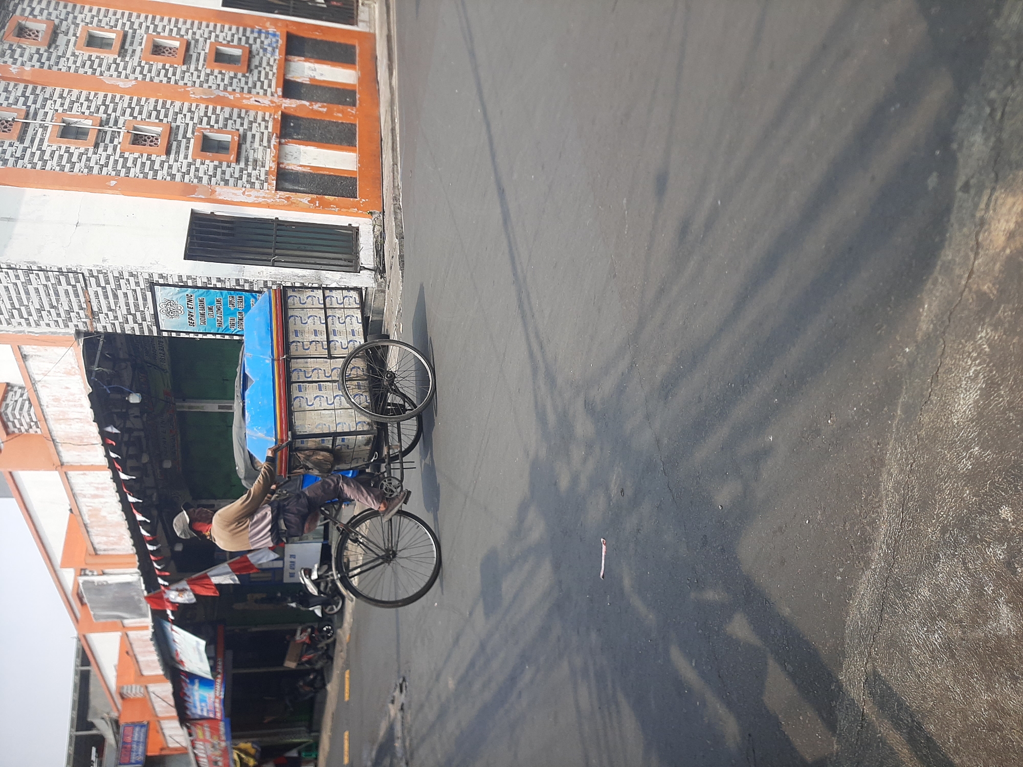 A person rides a pedicab, also known as a becak, down a street in Central Jakarta, Indonesia. The pedicab is laden with a blue plastic container and other items. The driver wears a light brown and pink long-sleeved shirt, dark pants, and a cap. The pedicab has two large rear wheels and a smaller front wheel. Buildings with orange and white striped facades and awnings line the street. The asphalt road shows patches of wear and tear, and shadows of the buildings and pedicab are cast across it. Several motorcycles are parked in front of one of the buildings.