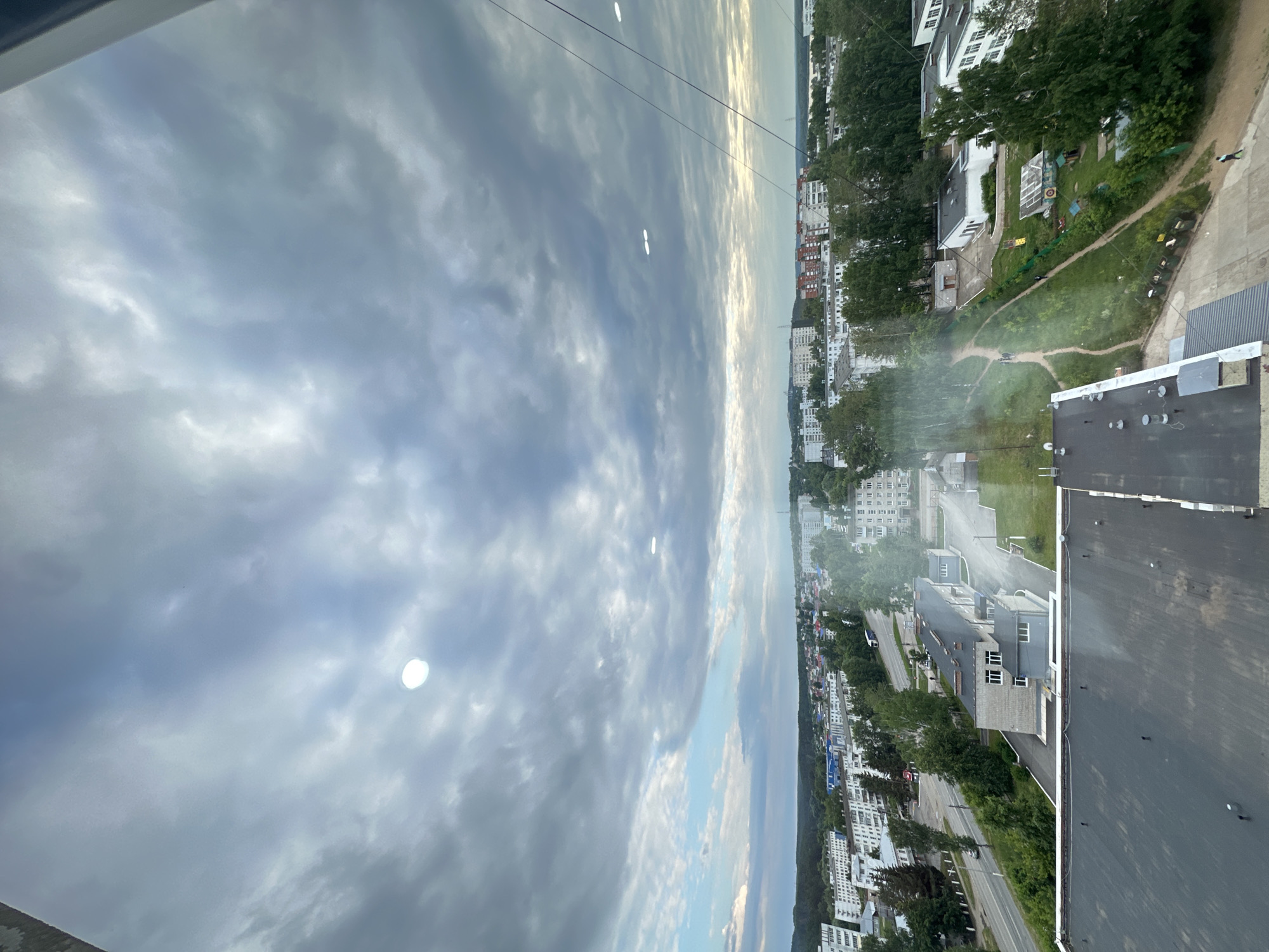 The image captures an aerial view of a city under a cloudy sky, likely taken from a high vantage point such as a tall building. The cityscape is characterized by numerous residential buildings, predominantly of a uniform, light-colored facade, typical of Soviet-era architecture, interspersed with green trees and smaller structures. Roads with visible traffic, including cars and a truck, are present, indicating an active urban environment. In the foreground, the dark, asphalted roof of a building is prominent, with several white vents or protrusions. The sky is heavily overcast with dramatic, textured clouds, but patches of lighter sky and hints of sunlight break through, creating a dynamic atmosphere. Two distinct, bright circular reflections, possibly from lights or glare on the window through which the photo was taken, are visible in the sky. The horizon line reveals a dense line of trees, suggesting the city is bordered by natural landscapes. A power line is also visible cutting across the upper right portion of the frame. The overall impression is one of a typical urban scene in a Russian city, possibly Chaikovsky, given the architectural style and the presence of apartment blocks.