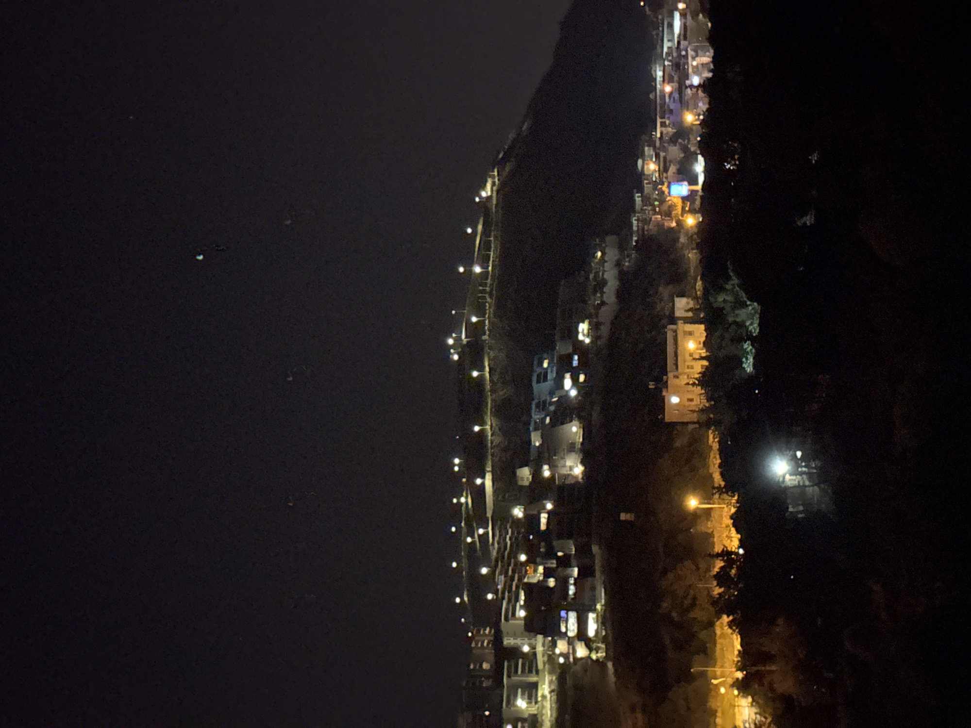 This nighttime image captures a panoramic view of Kuşadası, Turkey, from an elevated perspective, showcasing a developed urban landscape integrated with hilly terrain.
The upper third of the frame consists of a dark, clear night sky, with a few faint, indistinct specks of light visible. The majority of the image is occupied by dark hills and slopes, extensively populated with structures and illuminated infrastructure.
Numerous buildings, appearing as multi-story residential or commercial units, are constructed across the hillsides, characterized by varying architectural styles and illuminated windows and facades. These structures are scattered from the mid-ground, extending towards the upper slopes, and are particularly dense in the left and central portions of the mid-ground. Many buildings exhibit warm white and yellow interior lights, indicating occupancy.
A network of bright, spherical streetlights with white or warm yellow luminescence traces paths along the contours of the hills and roads, creating a distinct pattern of illumination that defines the urban layout. A prominent line of these streetlights ascends the main hill in the background.
In the lower mid-ground, a distinct, lighter-colored building with multiple illuminated windows stands out on a lower slope, suggesting a larger structure, possibly an institutional or significant residential property. Further to the right in the lower mid-ground, several brighter, more intense light sources, possibly from commercial establishments or signage, are visible amidst other structures.
Dark masses of trees and vegetation are present throughout the foreground and interspersed between buildings on the slopes, appearing as silhouettes against the ambient light. No individuals or specific human activities are discernible within the frame.