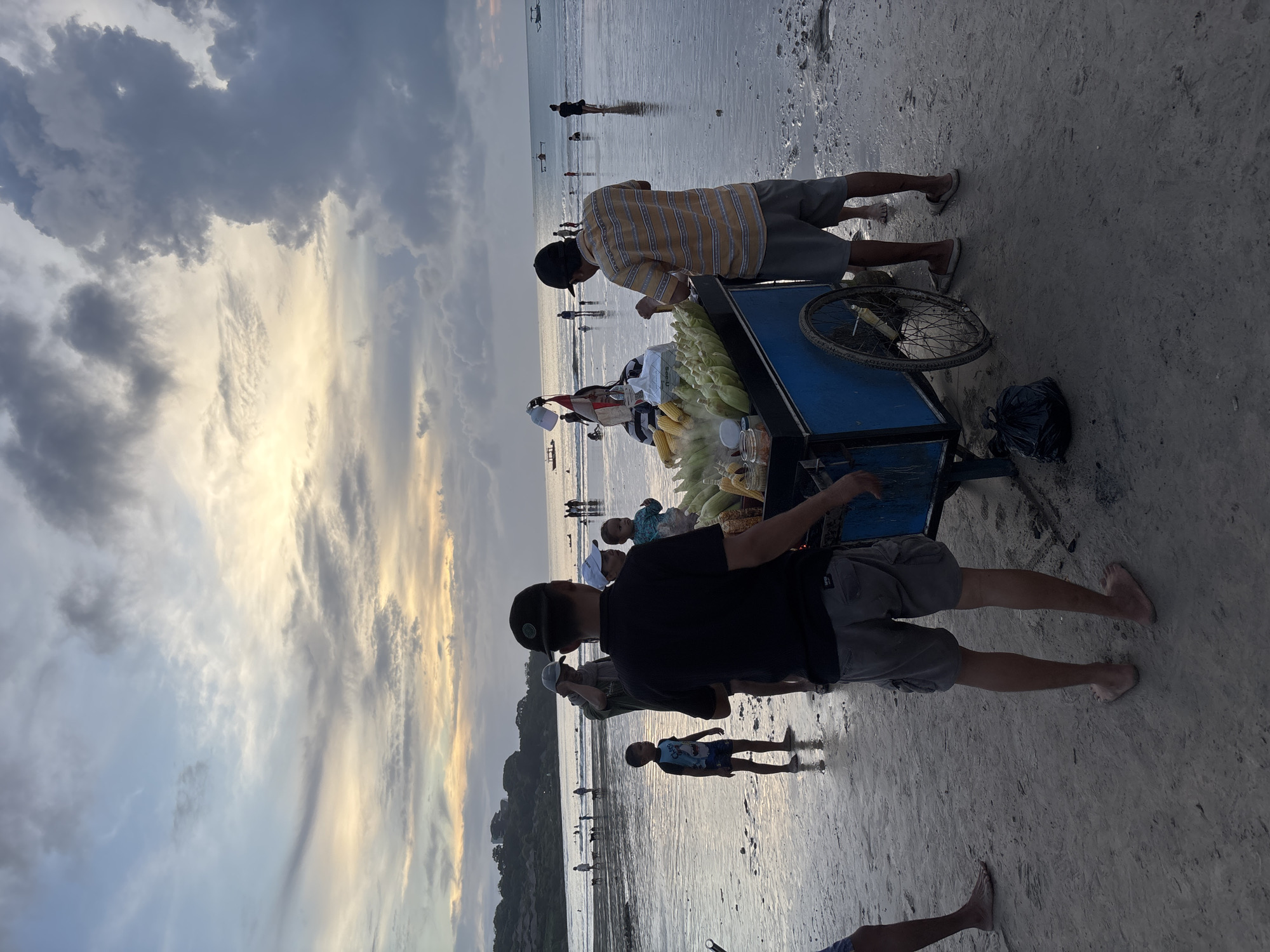 The image depicts an evening scene on a beach in Kuta Selatan, Indonesia, characterized by a dramatic sky featuring heavy clouds with warm, golden sunlight breaking through, suggesting sunset or twilight. The ground is wet sand, indicating the tide is out or waves have recently receded.
In the foreground, a blue pushcart with a bicycle wheel is set up on the sand. The cart is laden with numerous corn cobs, some fully husked and others partially, implying they are for sale. Two men are prominent near the cart. One man, dressed in a yellow and white striped shirt, dark shorts, and a dark cap, stands behind the cart, seemingly preparing or attending to the corn. The other man, wearing a black t-shirt, grey shorts, and a black cap, stands beside the cart with his back to the camera. Their presence and the cart's contents suggest they are street vendors.
Several other people are visible further down the beach and in the shallow water, appearing as silhouettes against the brighter horizon. Among them is a child in a light blue top and dark shorts standing near the foreground figures. In the far distance, more figures and small boats can be seen on the water. A black plastic bag rests on the sand next to the cart's wheel. The scene conveys a relaxed, bustling beach atmosphere during the evening. No visible text is discernible in the image.