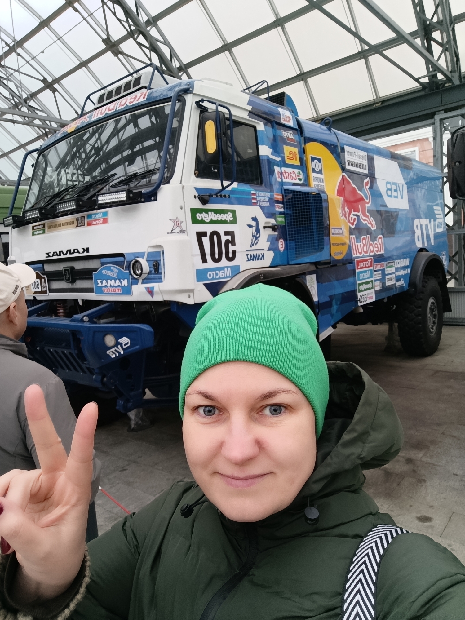 A selfie-style photograph captures a woman in the foreground making a peace sign, with a large, decorated rally truck in the background. The woman, wearing a bright green beanie and a dark green puffer jacket with a black and white striped strap, looks directly at the camera with a slight smile. Her left hand is raised, showing a peace sign.
Behind her, a formidable blue and white Kamaz rally truck is prominently displayed. The truck features numerous sponsor logos and racing insignia. The setting appears to be indoors, under a structure with a high glass and metal framed ceiling, possibly an exhibition hall or museum in Verkhnyaya Pyshma, Russia. Another person is partially visible on the left side of the frame, facing away from the camera.
Notable details on the truck include large "Red Bull" branding on the windshield visor and the side, a prominent number "201", and "KAMAZ" lettering on the grille. Many of the logos and texts are mirrored due to the selfie perspective. Visible text, including mirrored instances, includes: "KADRO", "KAMAZ", "KAMAZ-master", "201", "Red Bull", "VEB", "RTL", "Speed", "Castrol", "Knorr", "JATOT", "WECO", "OTBA", "WASCO", "SHEB", "TIEBHEBY". The truck is equipped with powerful LED light bars above the front bumper. The woman's expression is one of contentment or happiness.