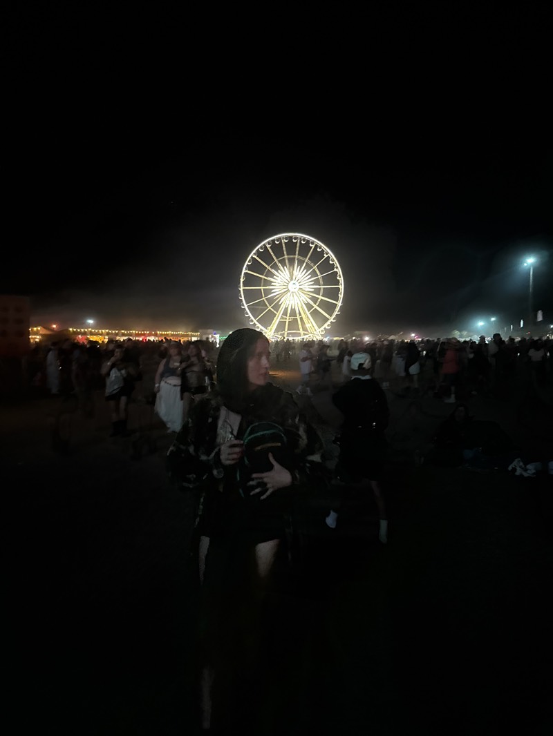 At night in Indio, United States, a crowd has gathered near a lit-up Ferris wheel. In the foreground, a woman holds a small backpack. Silhouettes of people fill the space between the woman and the Ferris wheel. Lights are visible in the distance.
