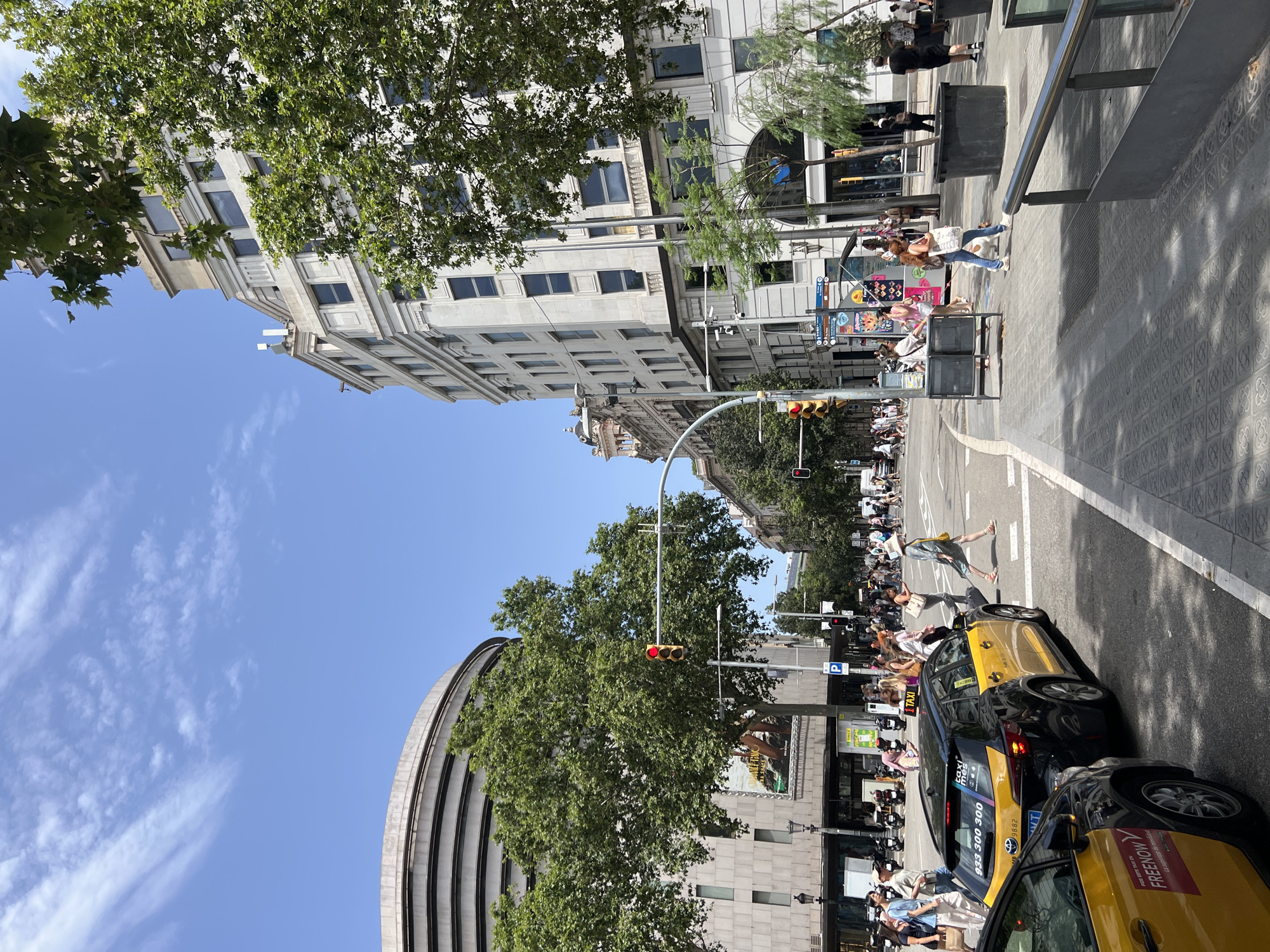 This is a ground-level, eye-level, outdoor, daytime photograph taken on a street in Barcelona, Spain. The image is oriented vertically, with the bottom of the frame facing upwards. A bright blue sky with wispy clouds is visible at the top.
The scene depicts a busy city street with several buildings, trees, and a significant amount of pedestrian and vehicular traffic. Several yellow taxis are prominently featured, some with branding that includes "TAXI" and phone numbers. One taxi has "FIREMOY" written on it. People are walking on the sidewalks and crossing the street. Some are carrying bags, and others appear to be interacting. A woman with an orange-brown top and jeans is walking a dog. Another woman in a yellow dress is crossing the street.
The architecture of the buildings suggests a European city, with ornate facades and balconies on some older structures, contrasted with more modern buildings. Lush green trees line the street, providing shade. Traffic lights are visible, with a red light illuminated on one. Several street signs and advertisements are present on the buildings and poles. The ground is paved with cobblestone-like patterns in some areas.
The time of day appears to be midday, given the bright sunlight casting shadows. The weather seems pleasant and warm. The overall atmosphere is one of a bustling urban environment.